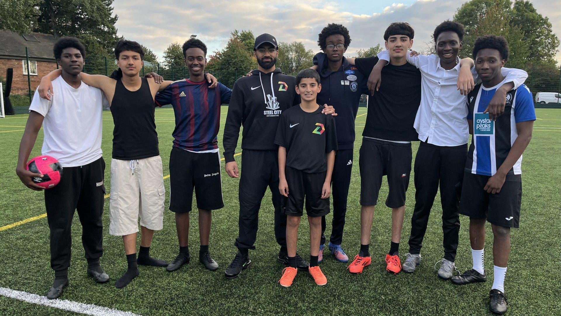 A group of teenage boys are standing in a line, with their arms around each other. They are all smiling at the camera. One on the far right holds a pink football. All are wearing football shirts, boots etc. In the centre is the project leader - who is older, wearing a baseball cap and hoody. They are stood in the middle of a football pitch.