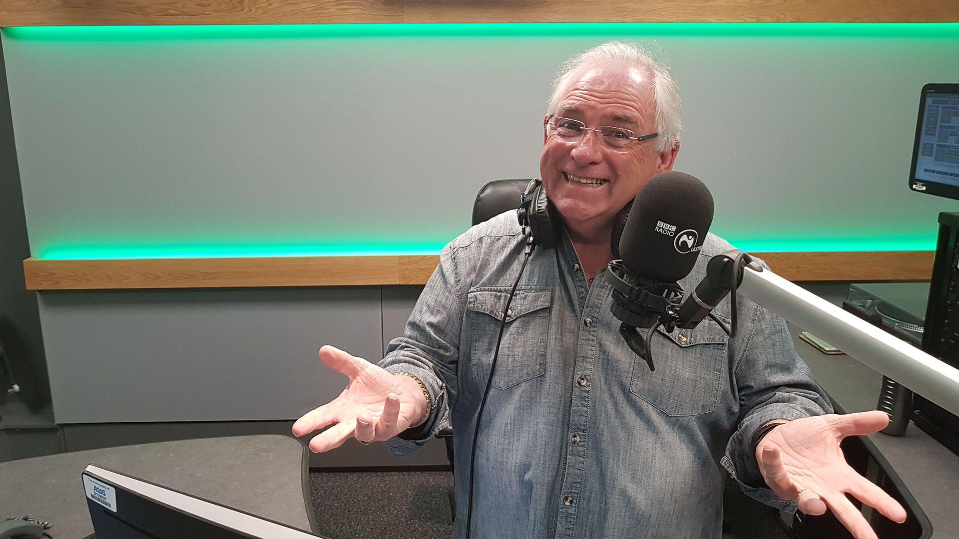 Hugo Duncan, smiling and wearing a grey shirt and glasses, sitting in a radio studio