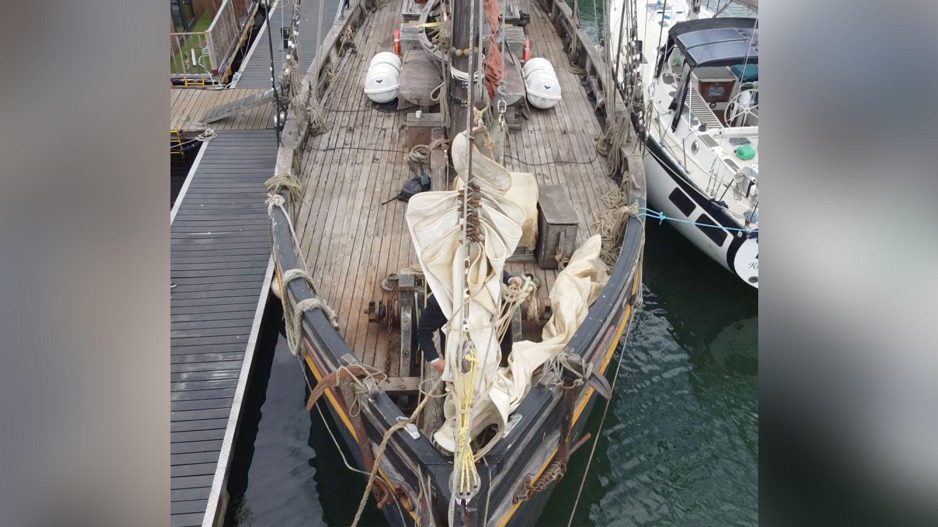 An aerial of the front of the Nordlys ship. The outside of the ship is painted black and the top deck is made from dark wood. The boat is moored on water and is sitting next to a white boat.