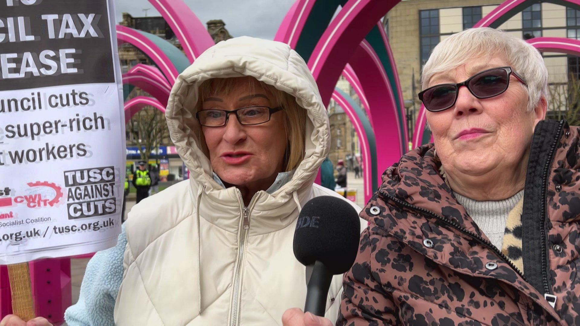 Two middle-aged women, both wearing coats, one wears normal glasses and the other sunglasses. One of them carries a placard protesting against council tax increases.