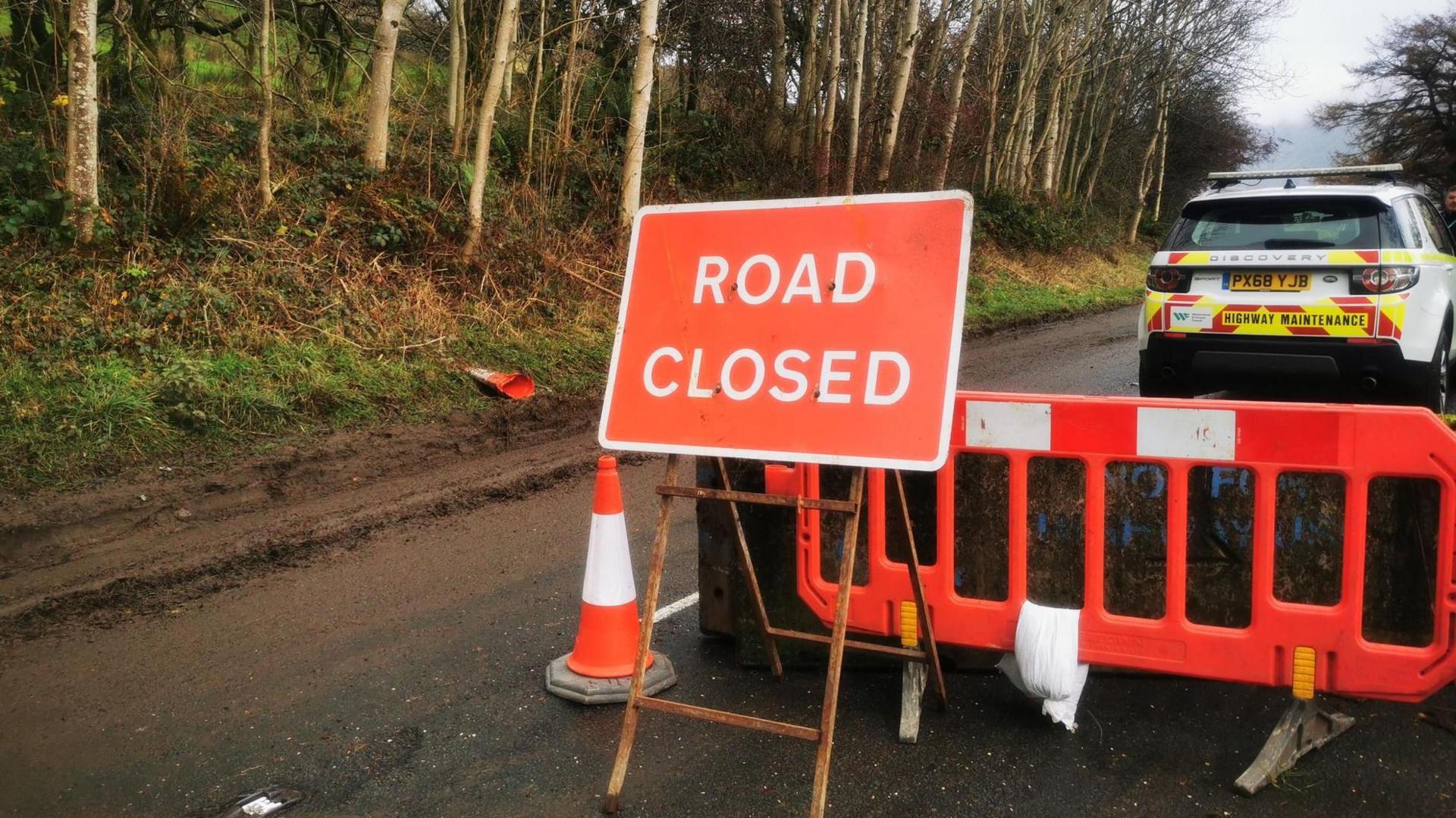 A red road closure sign stands in front of a plastic barrier and a concrete blockade. A highway maintenance vehicle is parked on the opposite side of the sign.