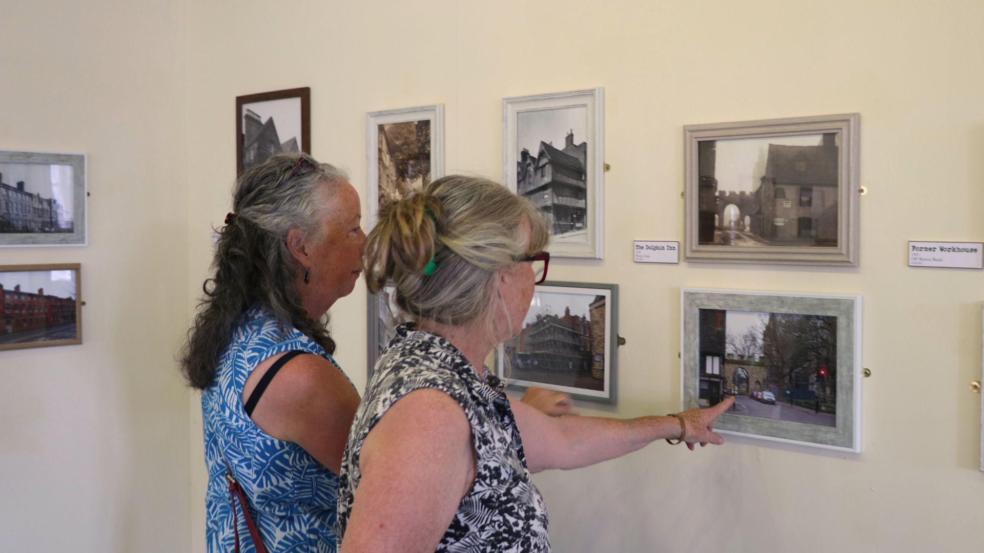 Two women are looking at photos, which are mounted on a wall in an exhibition, with archive photographs placed next to modern day re-creations of the same images.
