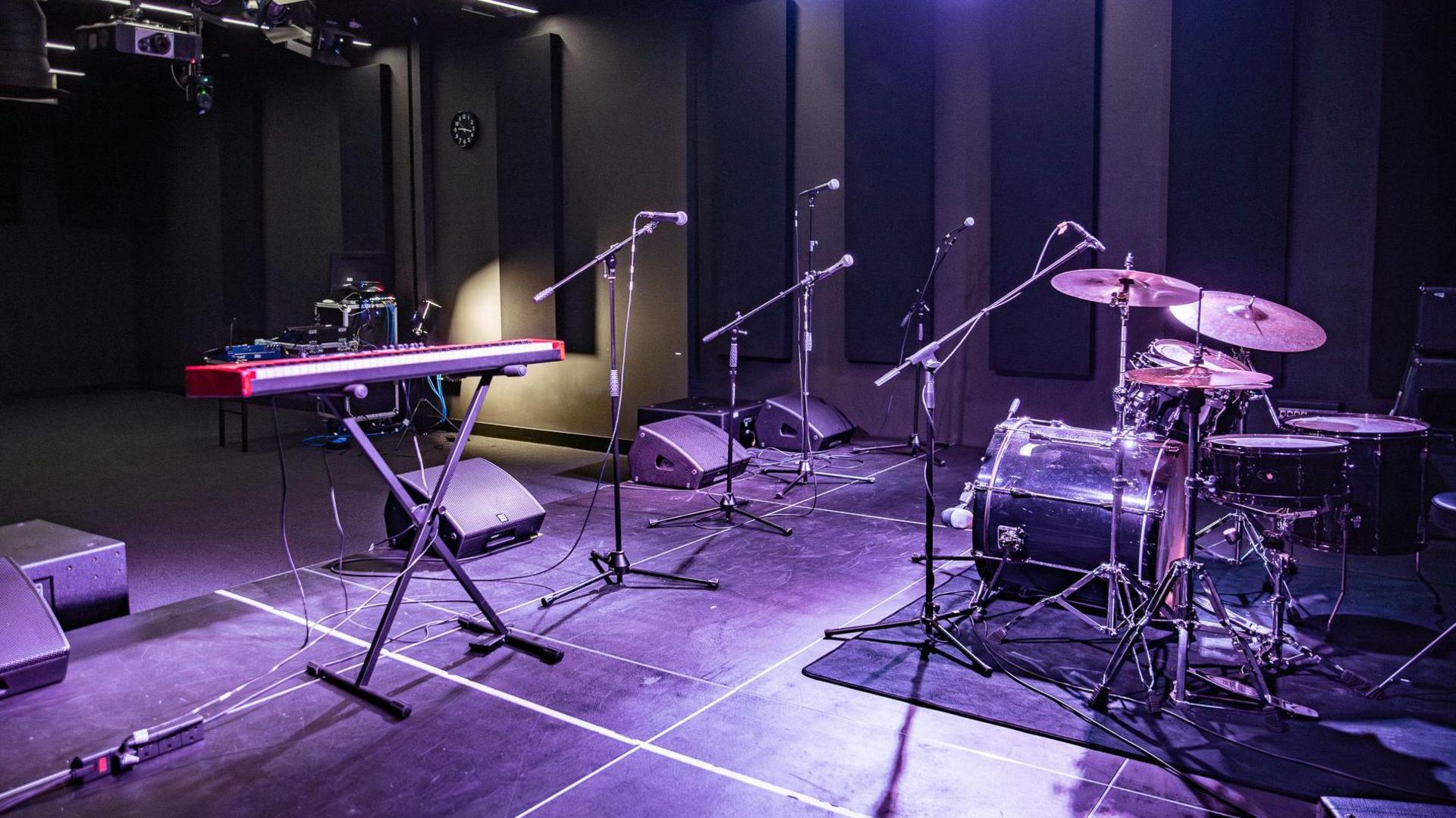A stage with an electric keyboard, microphones, drum kit and speakers with professional lighting. 