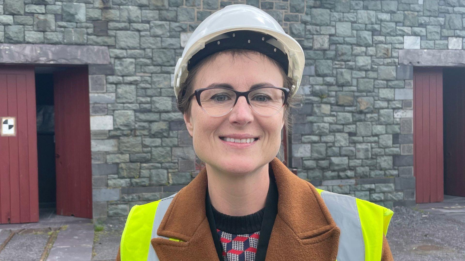 Llun o Helen Goddard yn sefyll ar safle'r amgueddfa. Maen gwisgo helmed gwyn a fest 'high-vis'. Adeilad cerrig yn y cefndir.