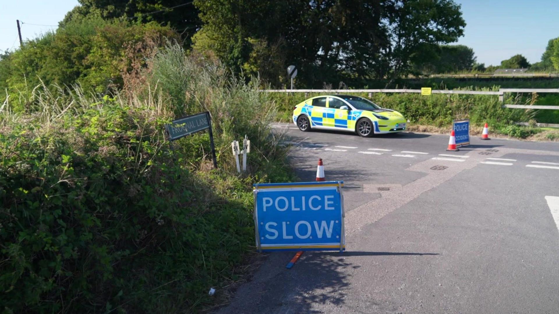 A police car sits still at the scene with a blue Police Slow sign