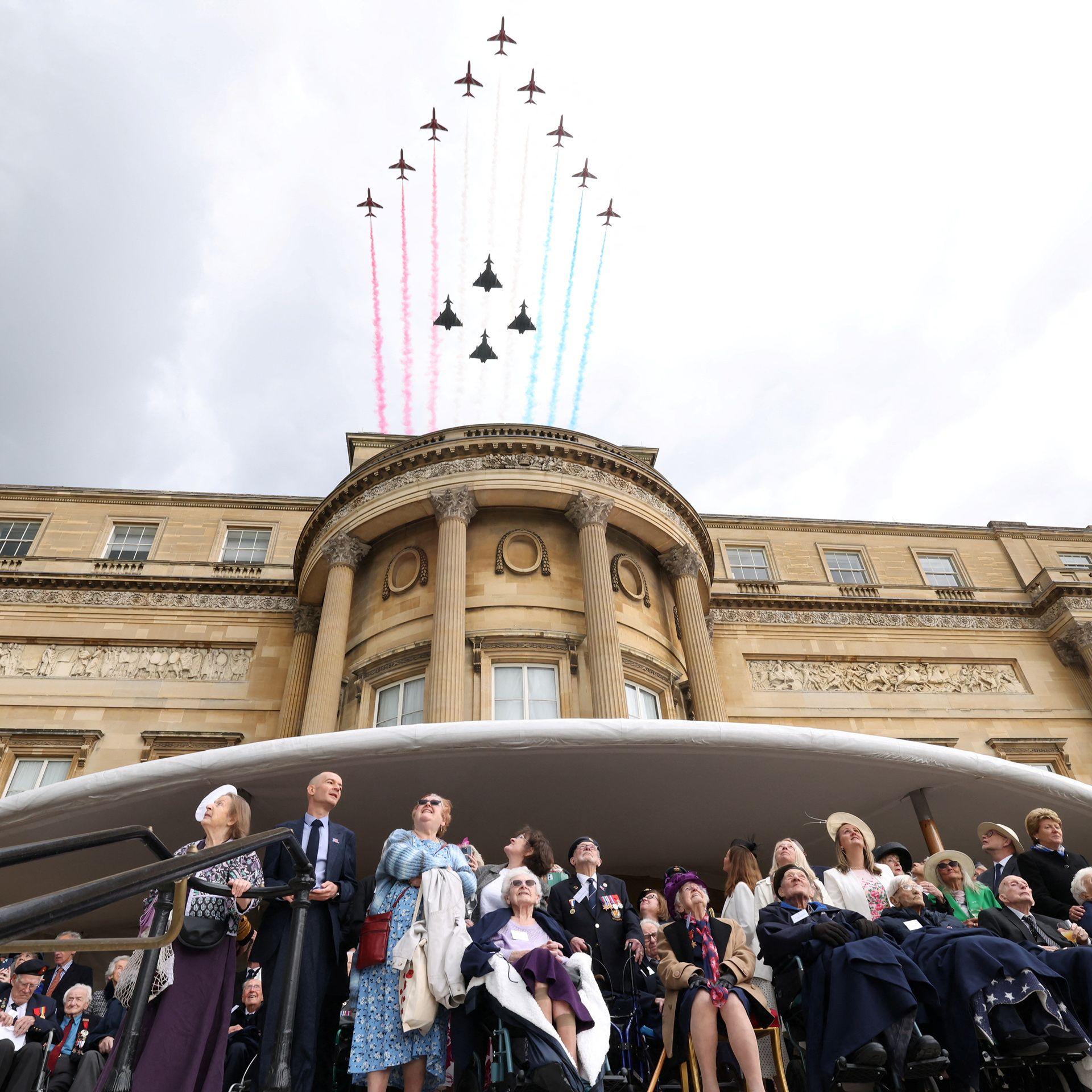 Red Arrows deploy coloured smoke as they fly over Buckingham Palace with a group of veterans all looking up to the sky below. It's a moody, grey cloudy day.