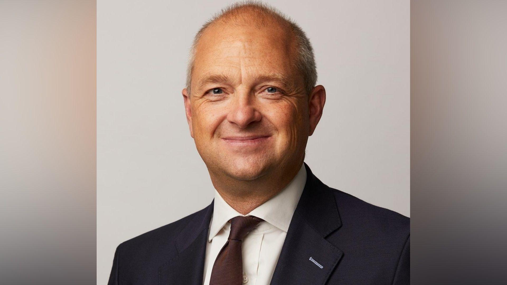 Jerome Mayhew looks towards the camera and smiles, wearing a dark suit, white shirt and tie