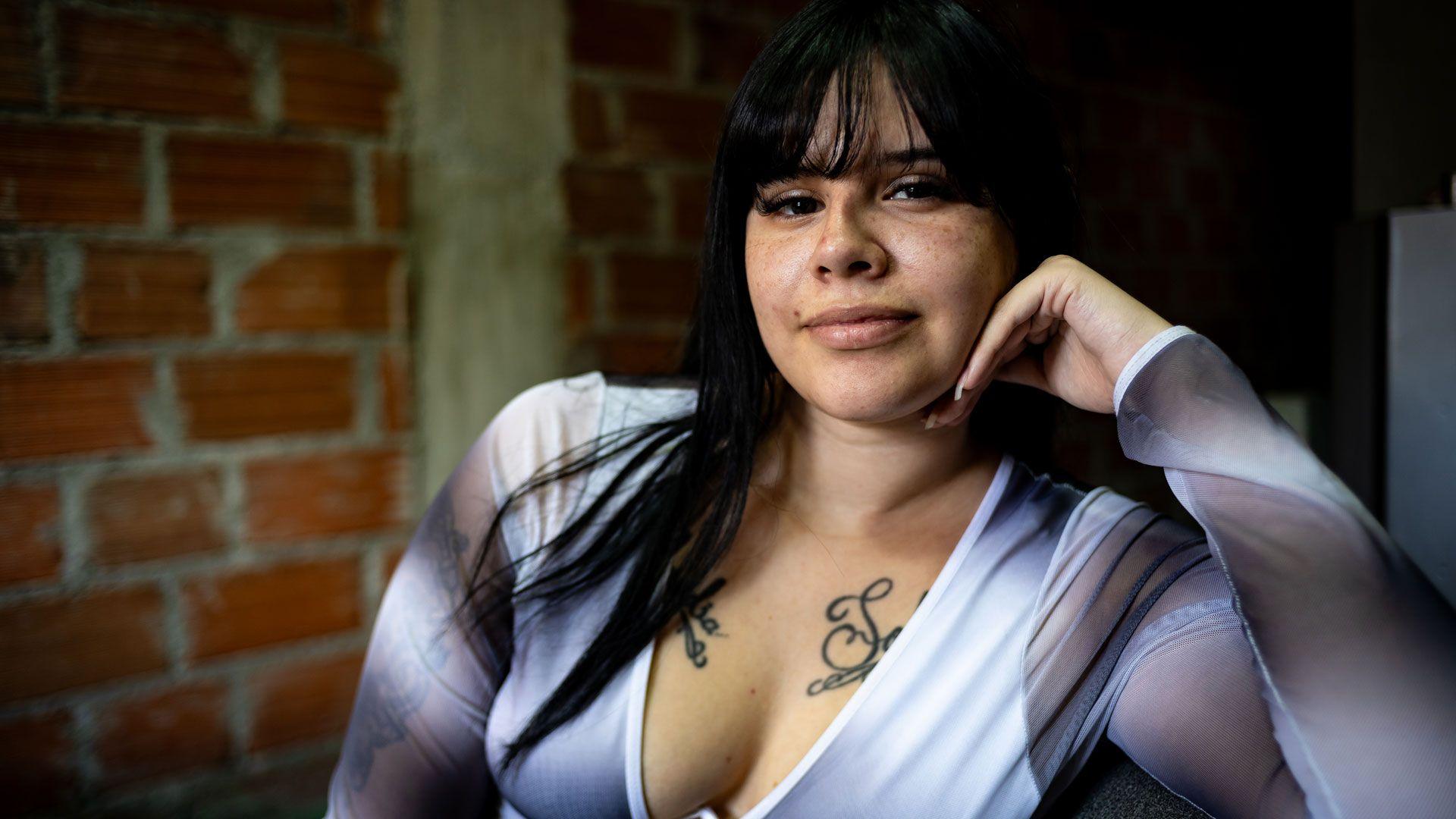 A young woman with long black hair in a low-cut top with tattoos on her chest looks into the camera, with a slight smile on her face.