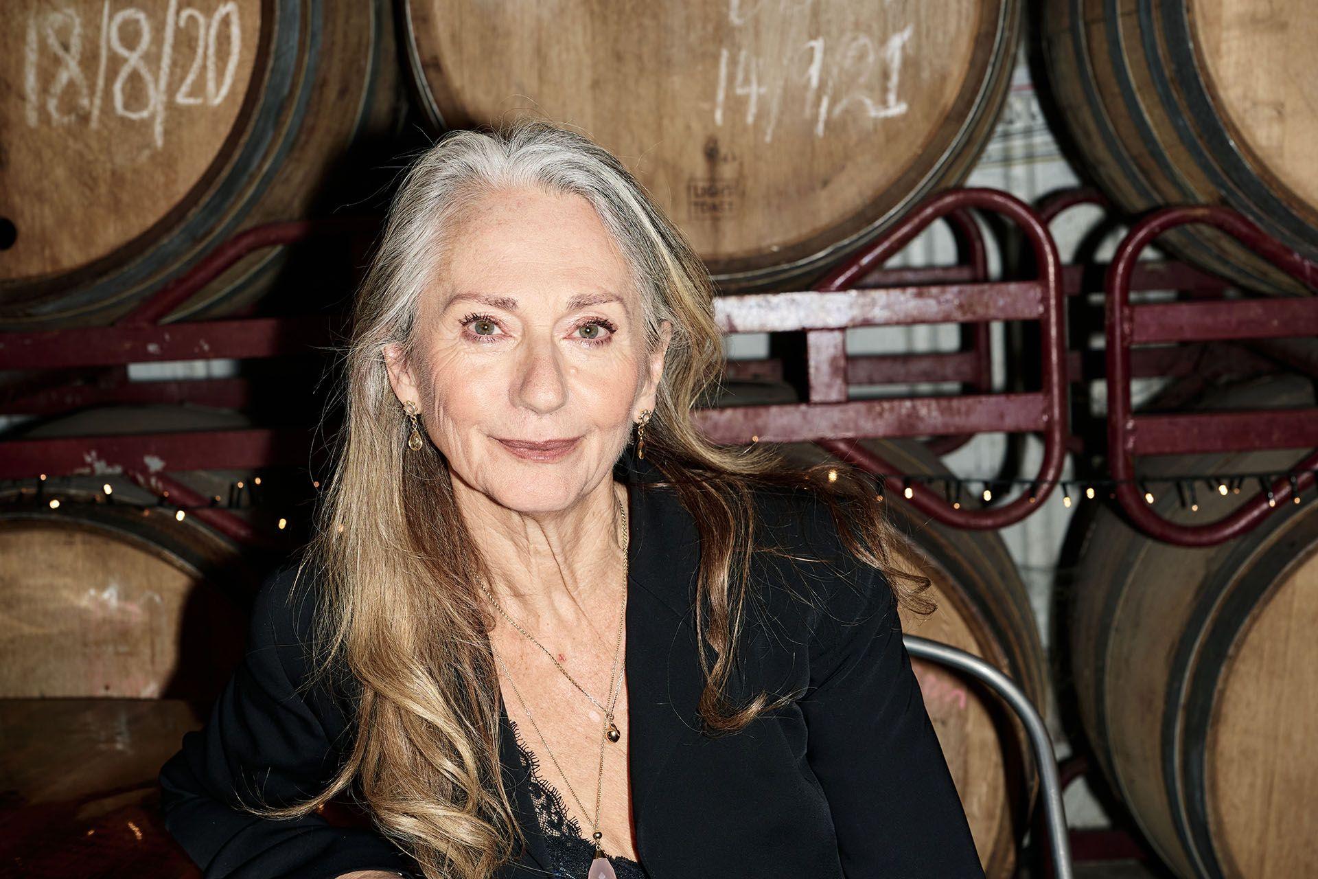 rachel joyce sits in wront of a series of barrels. she has brown hair that is grey at the roots and wears a black jacket