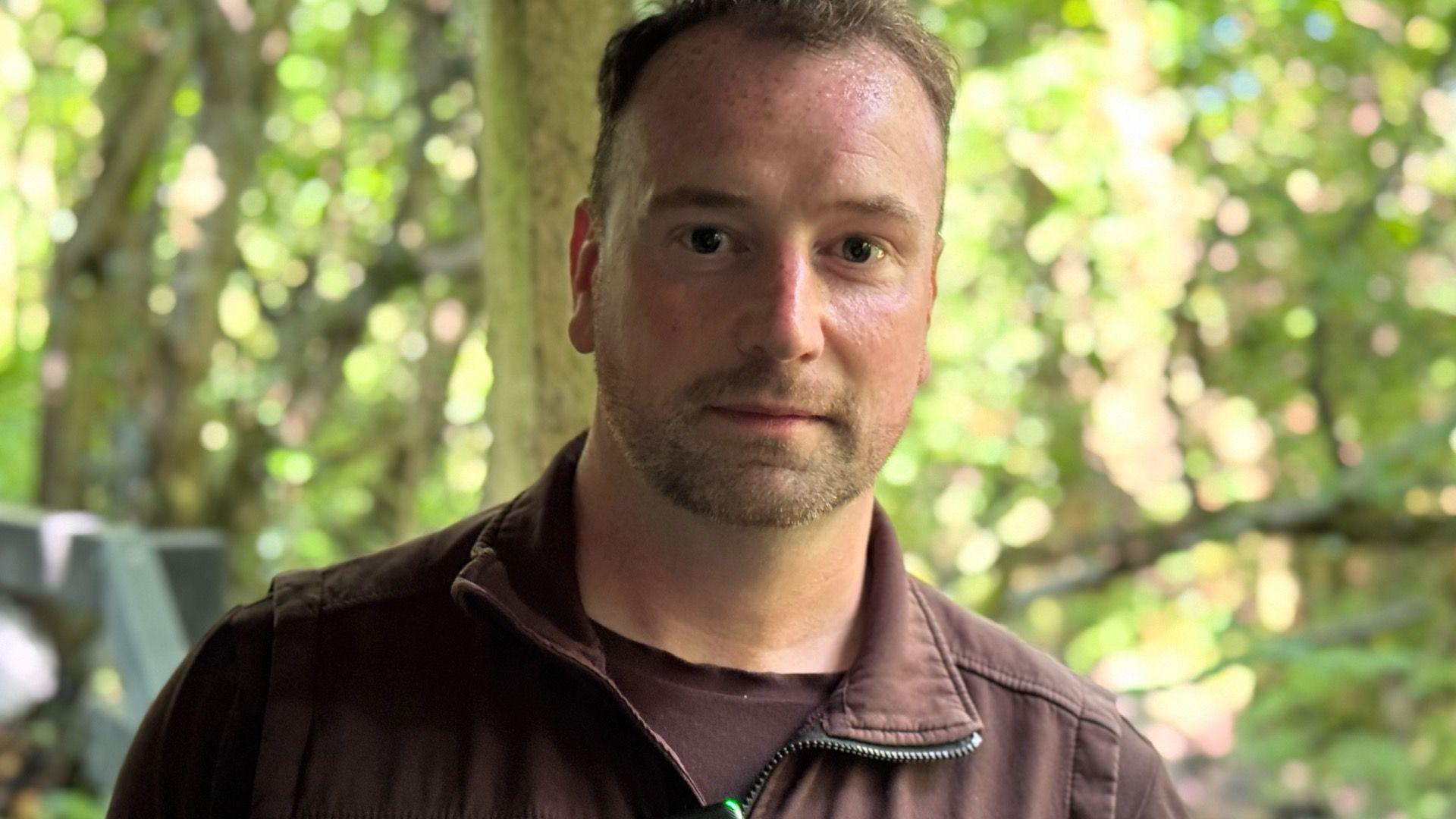 Peter Mitchell has short light brown hair and a beard. he is wearing a brown zip up gilet over a brown t-shirt and is standing in a wooded area.