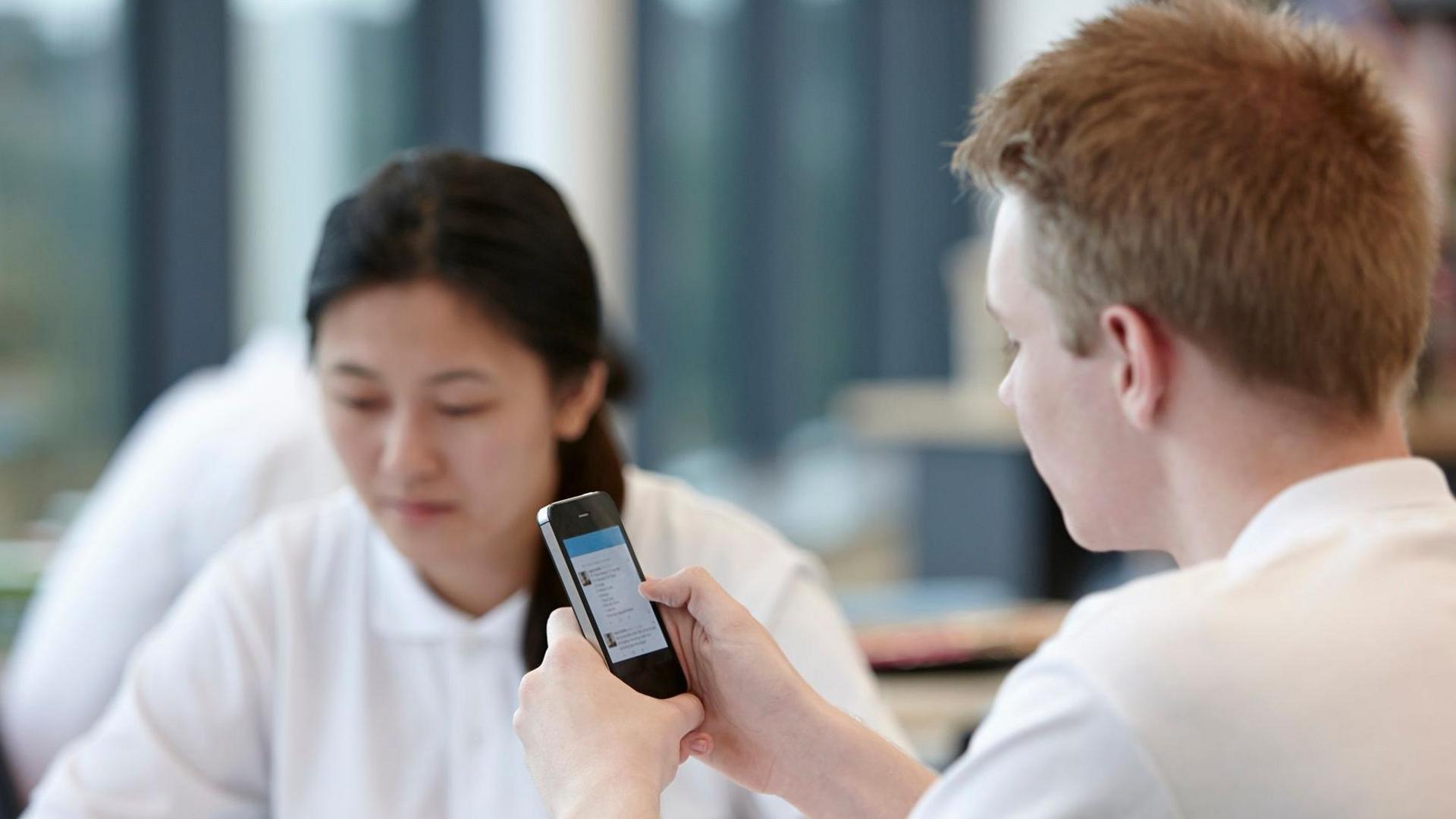 Boy dressed in school uniform with mousy sat in classroom looking at his phone. In the background, a girl with dark hair is looking down at her work.
