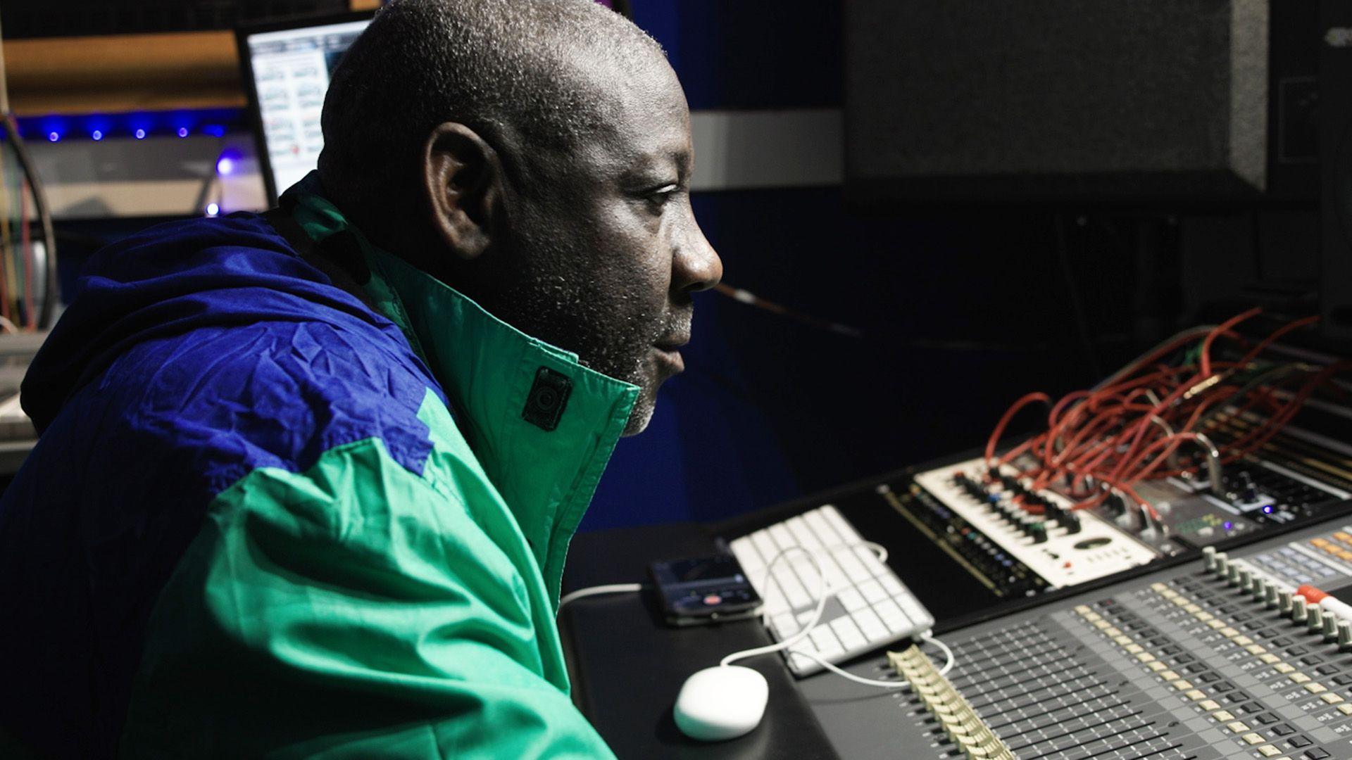 A man wearing a blue and green jacket is sitting in front of a music studio console