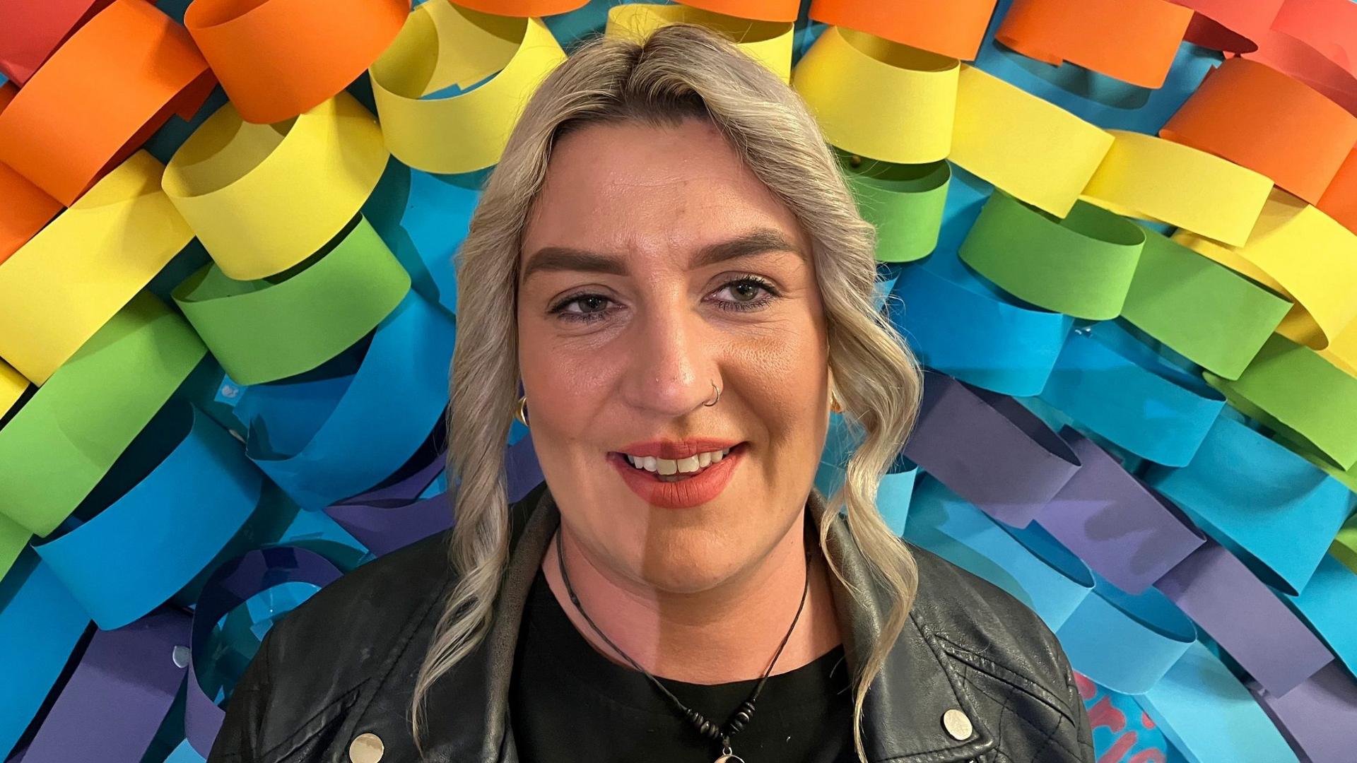 Trisha O'Connor, who is attending the class to help her son, stands with a smile, wearing a black leather jacket over a black t-shirt with a circular necklace. She has curly blonde hair, wears a nose ring, and is standing in front of a colourful wall pattern.