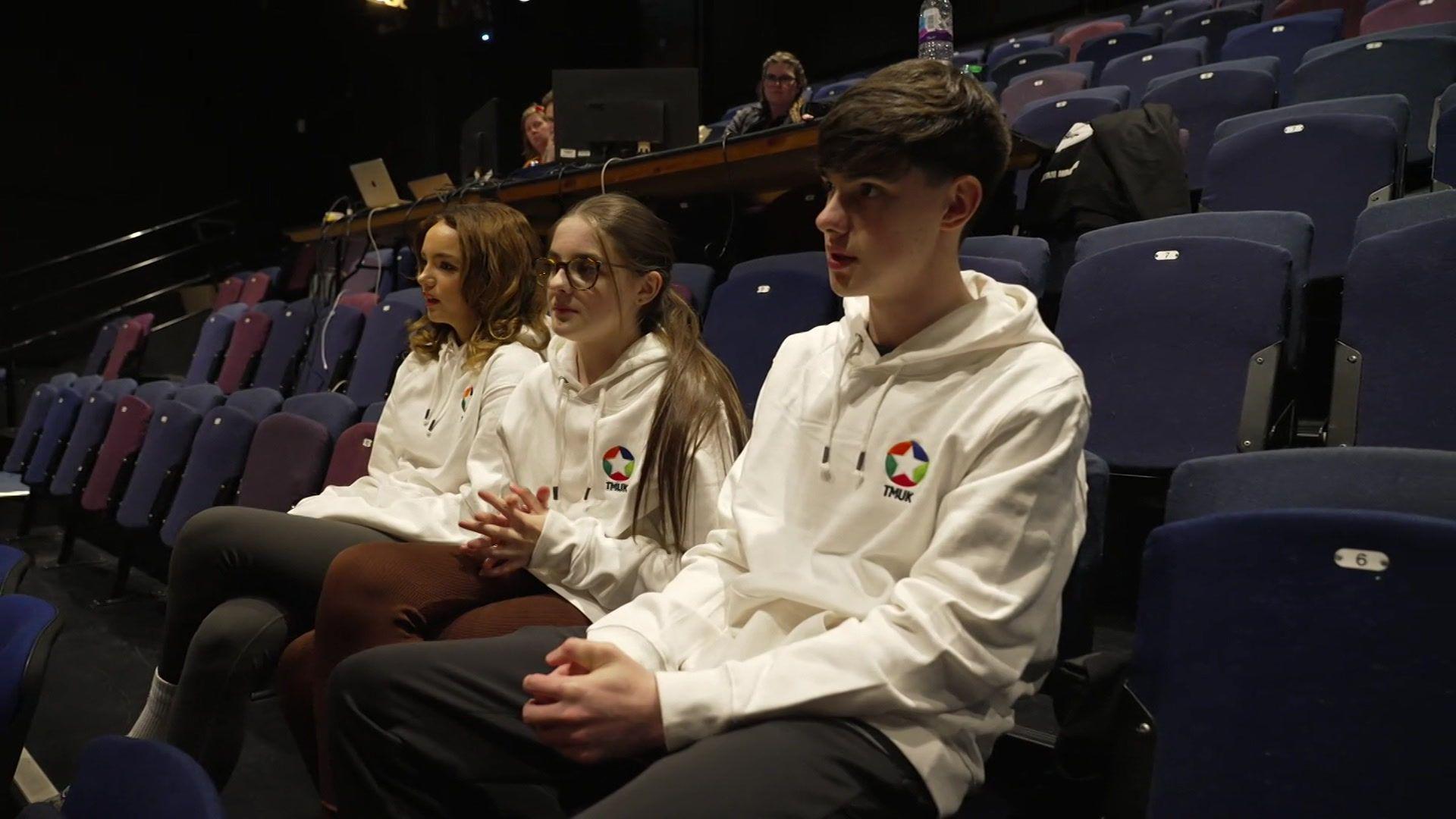 Bella, Cerys and Aaron at the rehearsal, sitting on blue theatre seats and wearing matching white hoodies displaying the star-shaped logo of Time-Matters UK