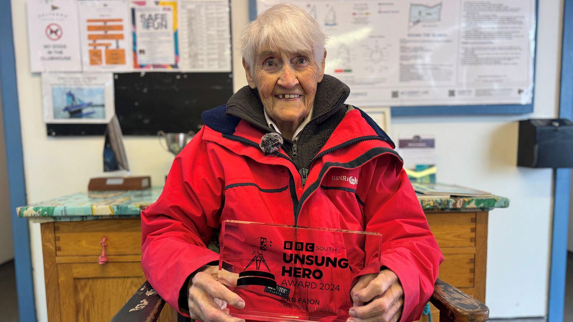 Jean Paton sits down wearing a red waterproof coat holding her BBC South Unsung Hero award.