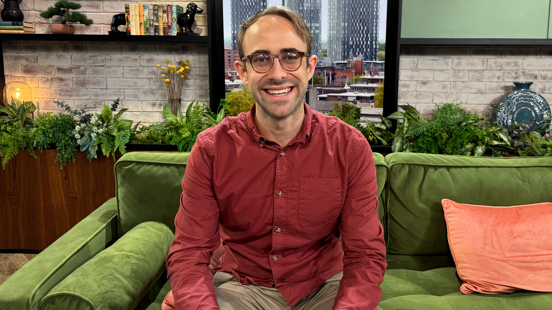 Harry Kind, wearing a red shirt and glasses, sitting on the green sofa in the Morning Live studio, with brick walls, bookshelves and a view of Manchester city centre visible behind him.