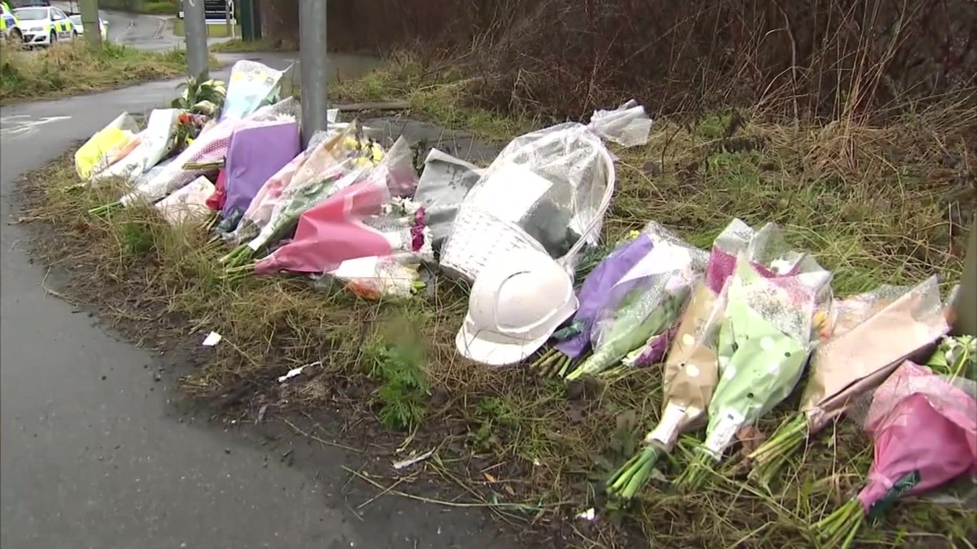 A row of flower bouquets, gifts and helmets placed in memory of the plant collapse victims. It is an overcast day. Police cars can be seen in the distance.