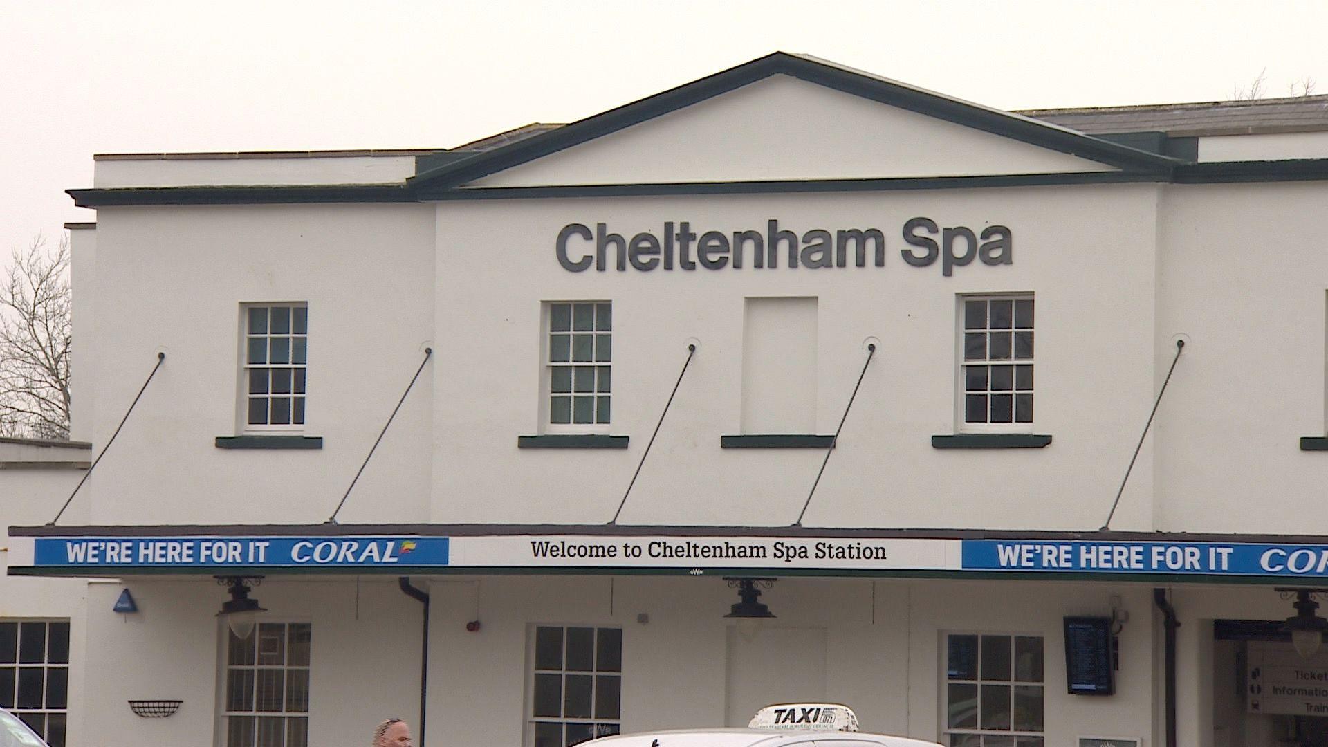 The exterior of Cheltenham Spa railway station, a two-storey white rendered building with betting shop advertisements. It has a large sign reading the name of the station and the top of a taxi can be seen waiting outside.
