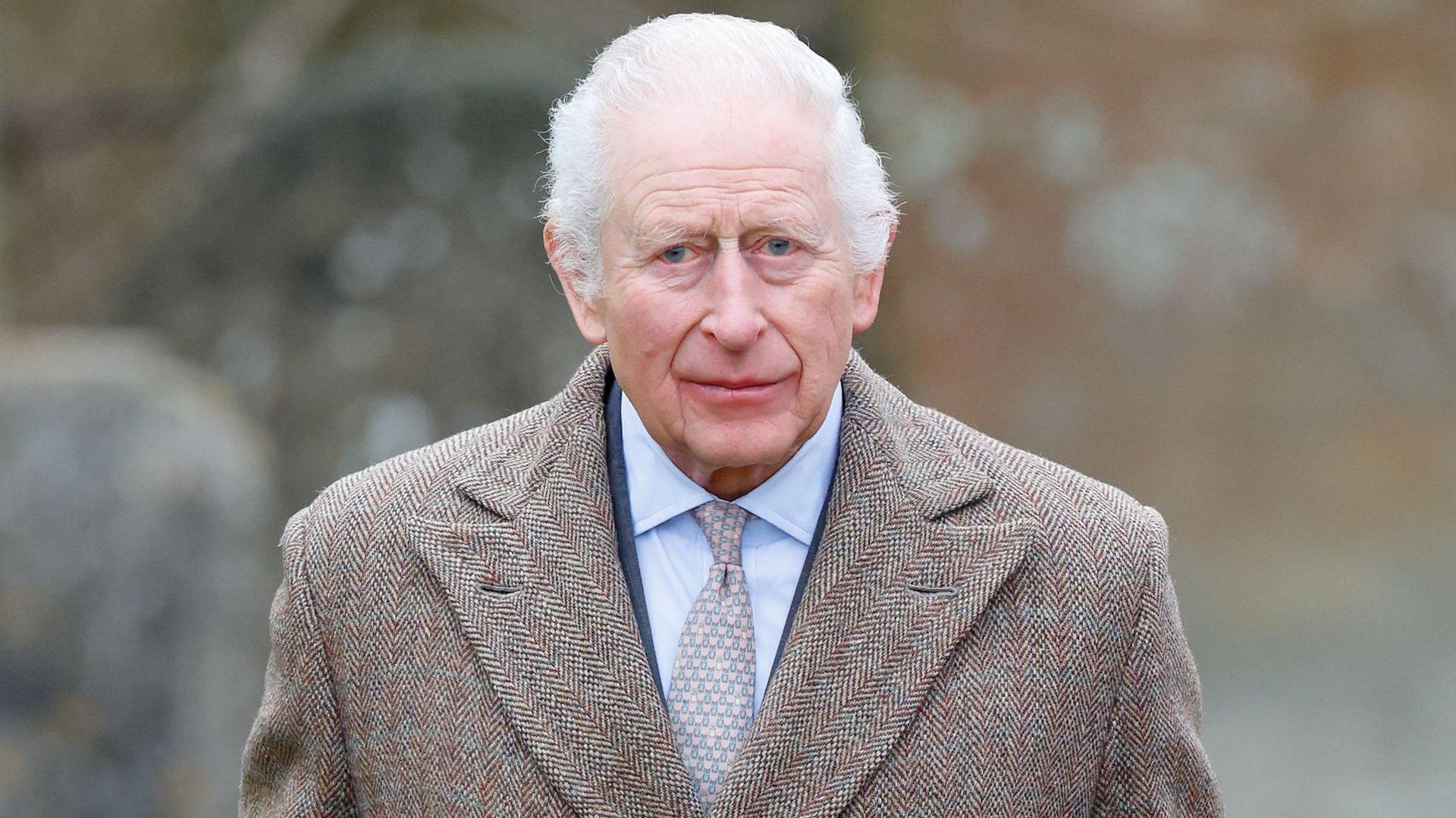 King Charles III attends the Sunday service at the Church of St Mary the Virgin, close to the Sandringham Estate. The King is wearing a brown tweed coat, navy suit and a lightly decorated tie. He is looking towards the camera lens from a distance