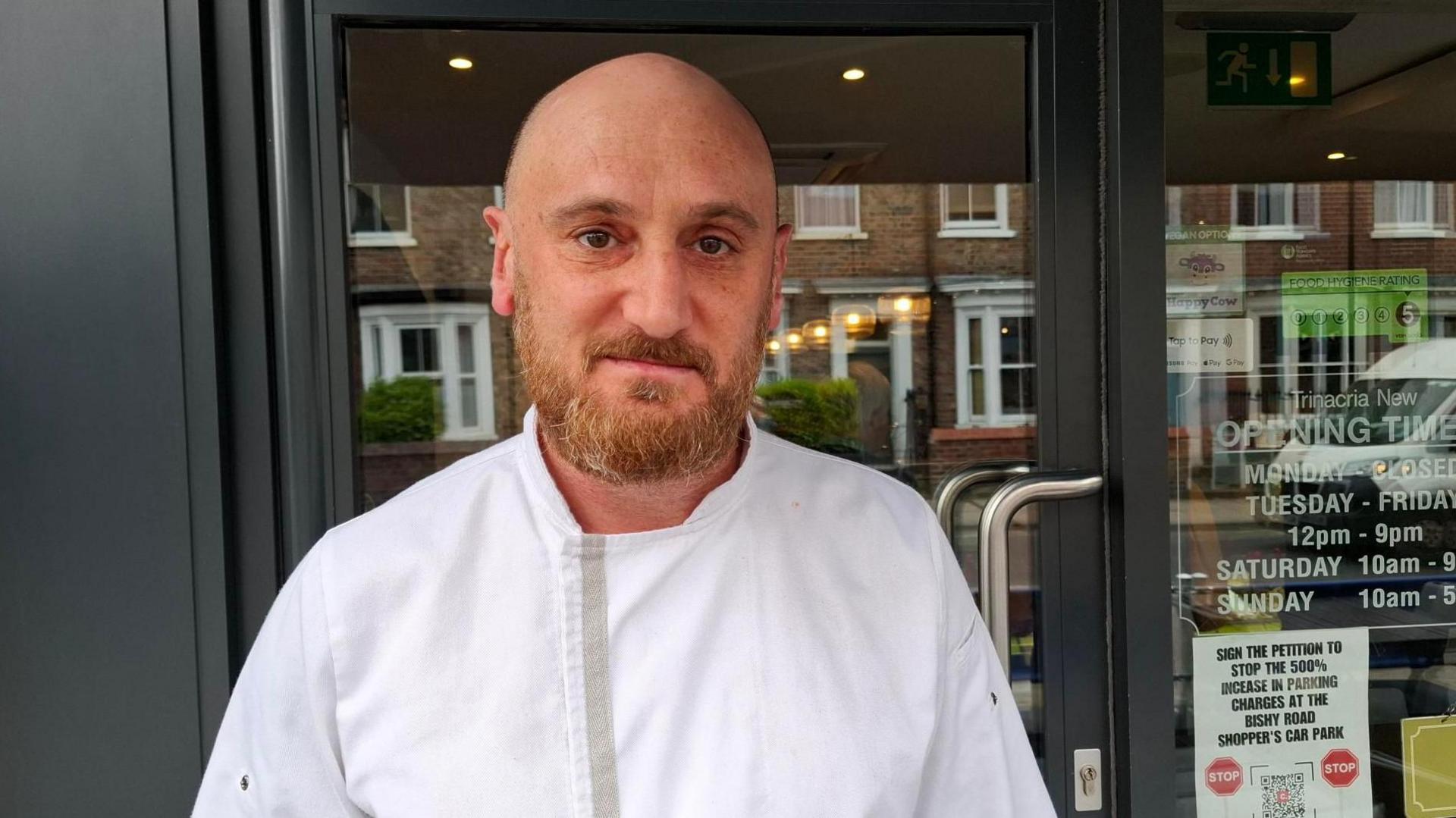 Beppe Lombardo has a bald head and a ginger beard. He is wearing a chef's uniform and is standing outside a restaurant door.