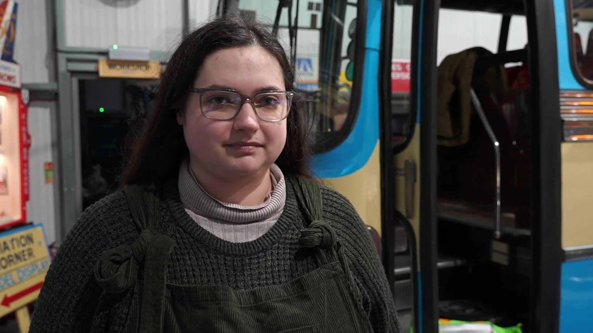 Kethry Heather looks slightly away from the camera, half smiling. She stands in front of a bus in the museum. She has long black hair and is wearing a dark grey jumper with a lighter grey turtle neck top underneath. She also has glasses.