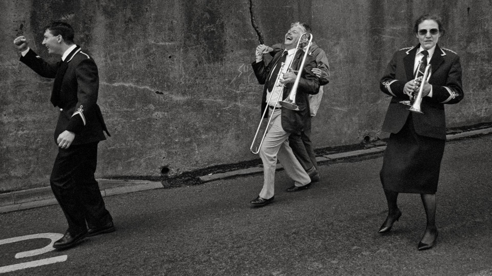 Four people who are part of a brass band are walking down the street. One man is carrying a trombone and a woman is holding a trumpet. Two of the men are laughing, one has his head thrown back and is holding the hand of someone behind him. The man at the front of the four, who is wearing a brass band uniform, has his right fist raised.