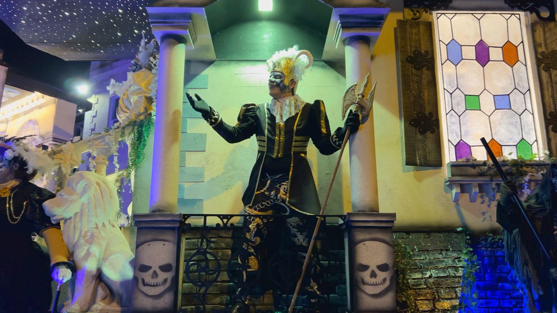 A performer dressed in costume. They are wearing a black and gold gown with a white wig. They are carrying a gold staff in one hand. They are standing on a cart which appears to be decorated for halloween, with two skulls on the side.