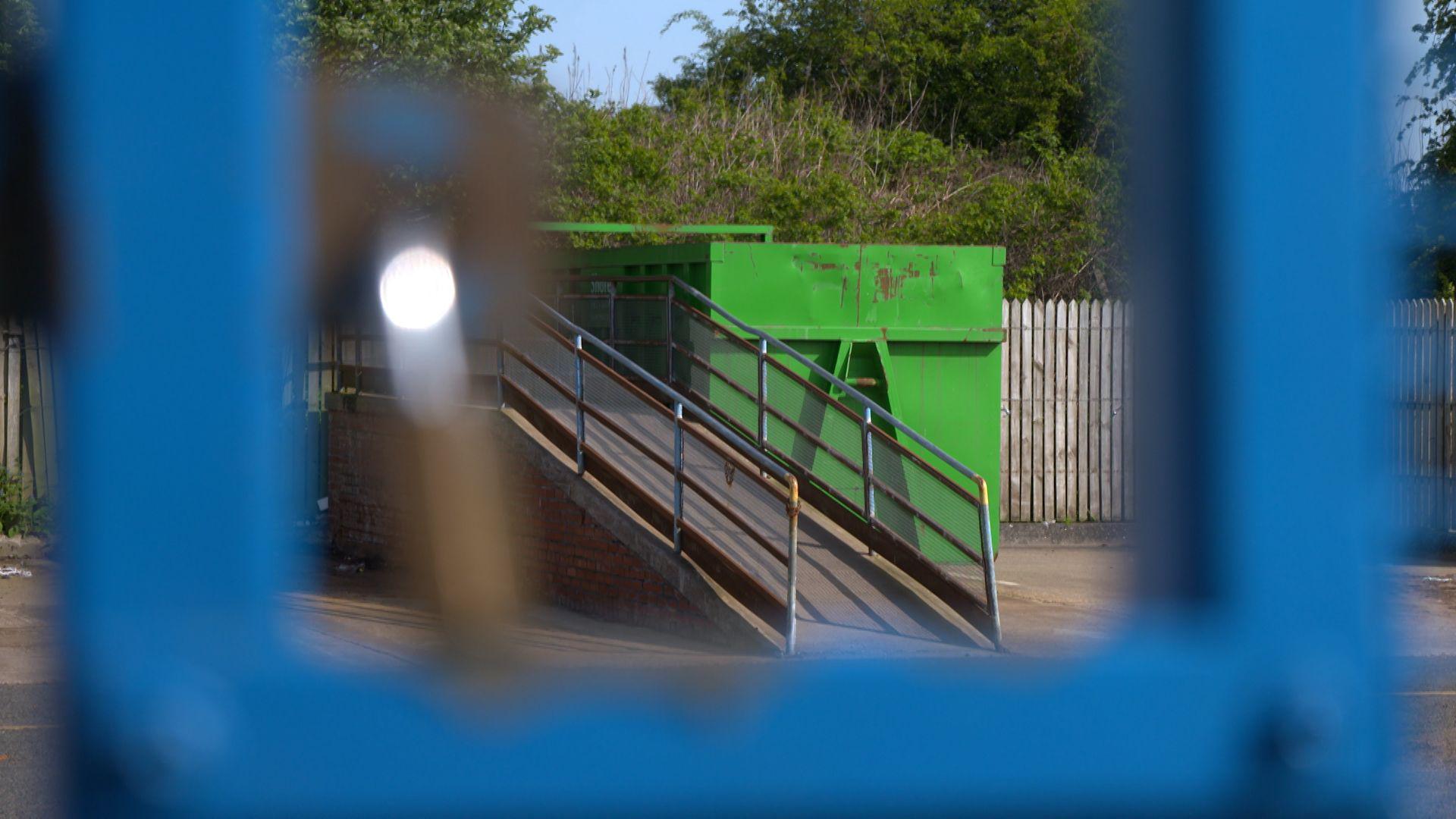 A green skip at a household recycling centre viewed through blue railings