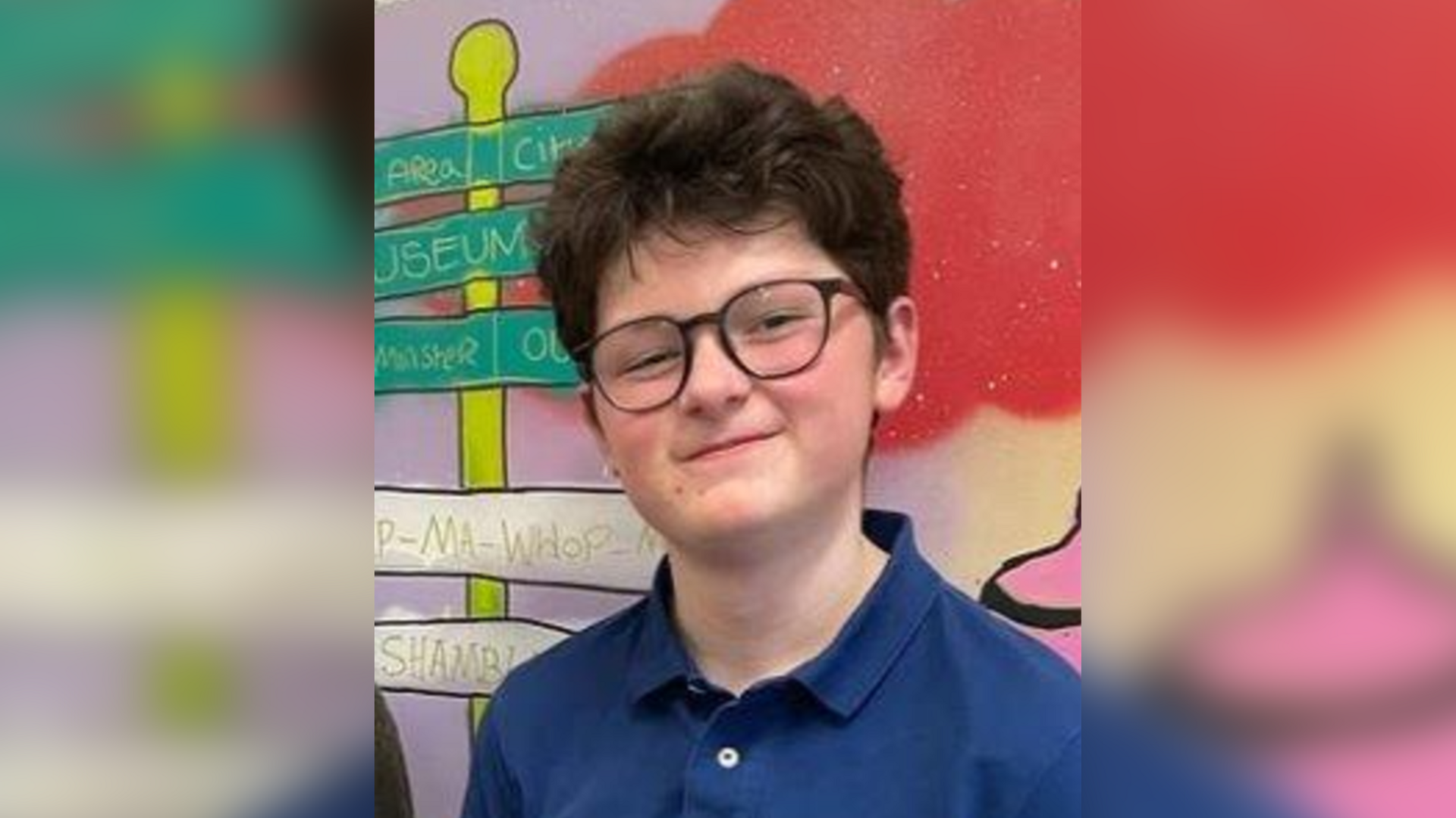A teenage boy wearing glasses with brown hair smiles at the camera. He is standing in front of a wall painted with colourful artwork.