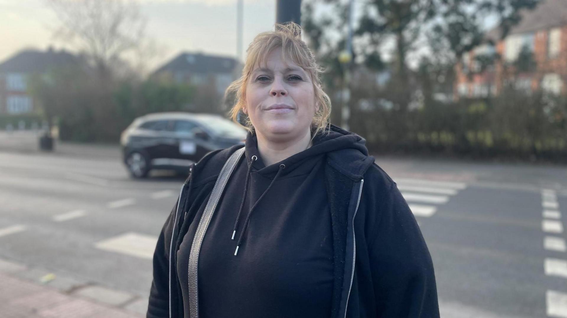 A woman with fair hair in a pony tail and a black hoodie stands looking cross in front of a zebra crossing