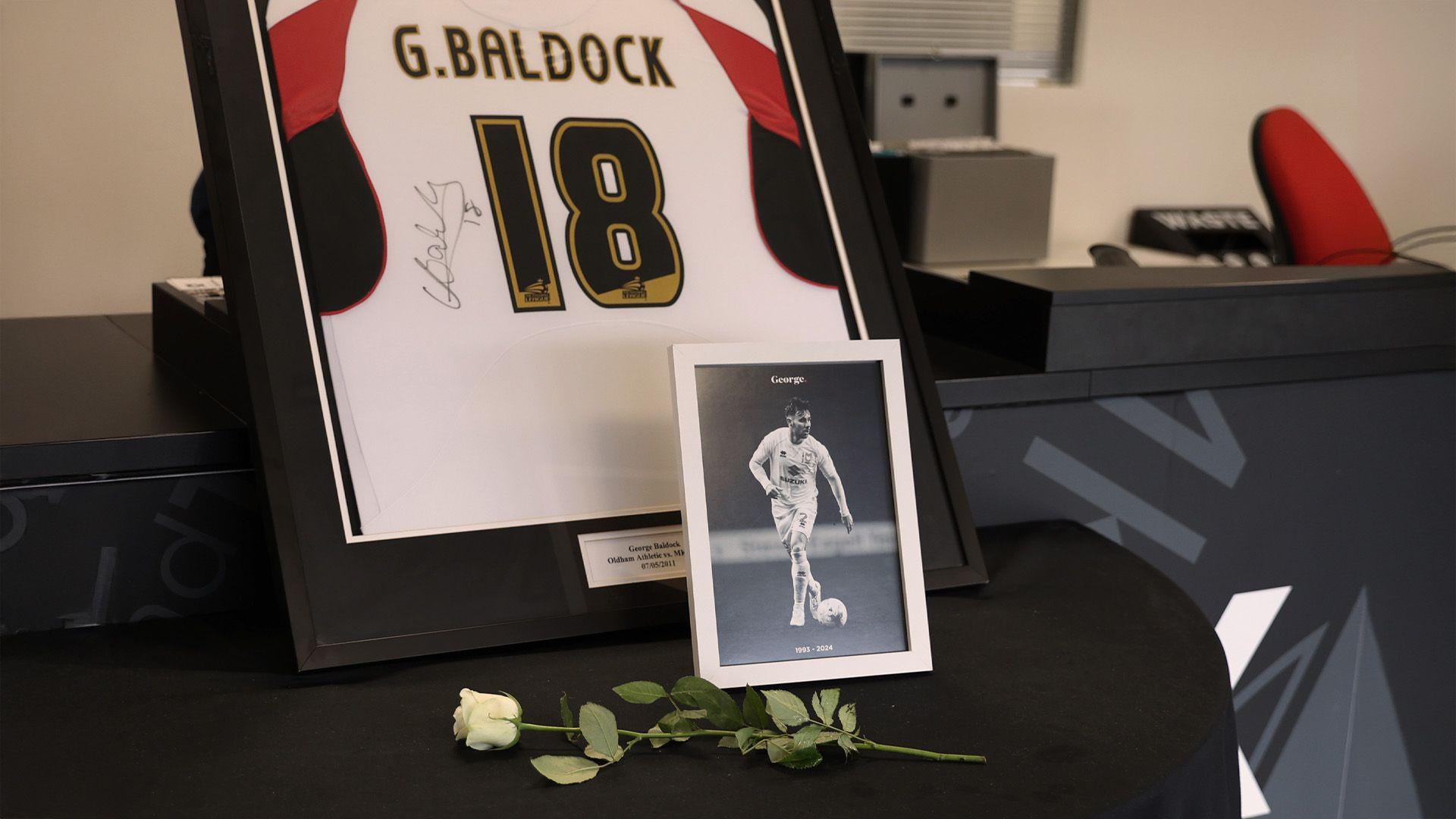 A framed black and white photo of George Baldock sits next to a white rose and his framed and signed football kit which features the number 18.