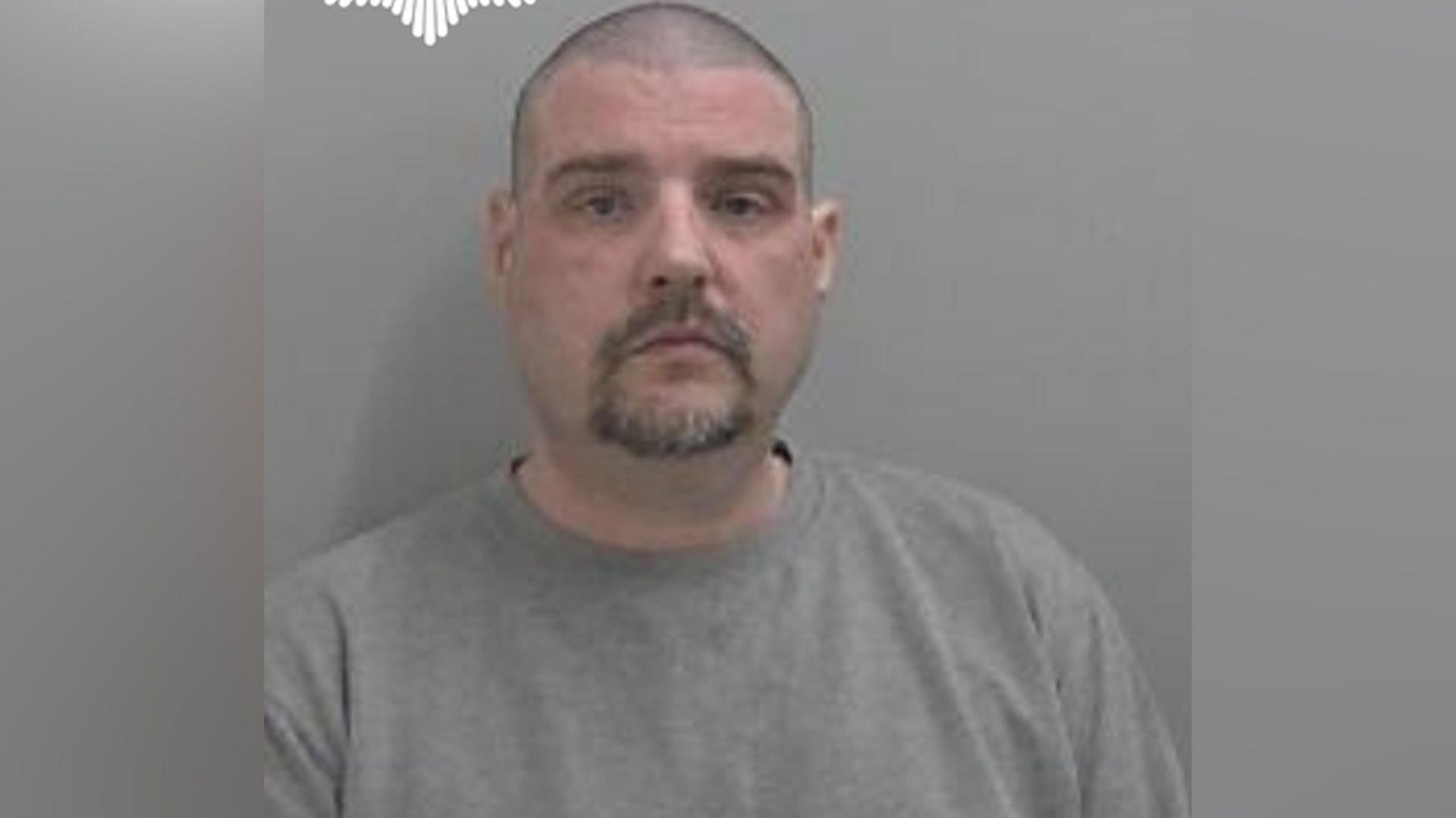 Chris Hemming pictured in a mugshot. He has a blank expression and wears a grey jumper. He has dark stubbly hair and a dark beard.