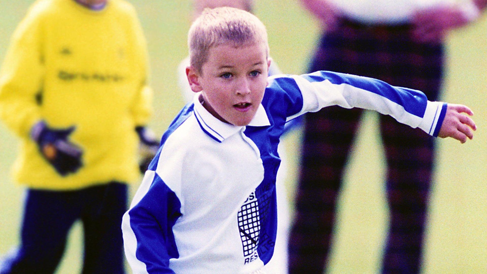 Harry Kane is seen playing for Ridgeway Rovers as a child. The archive image shows him in his blue and white kit while people watch on behind him.