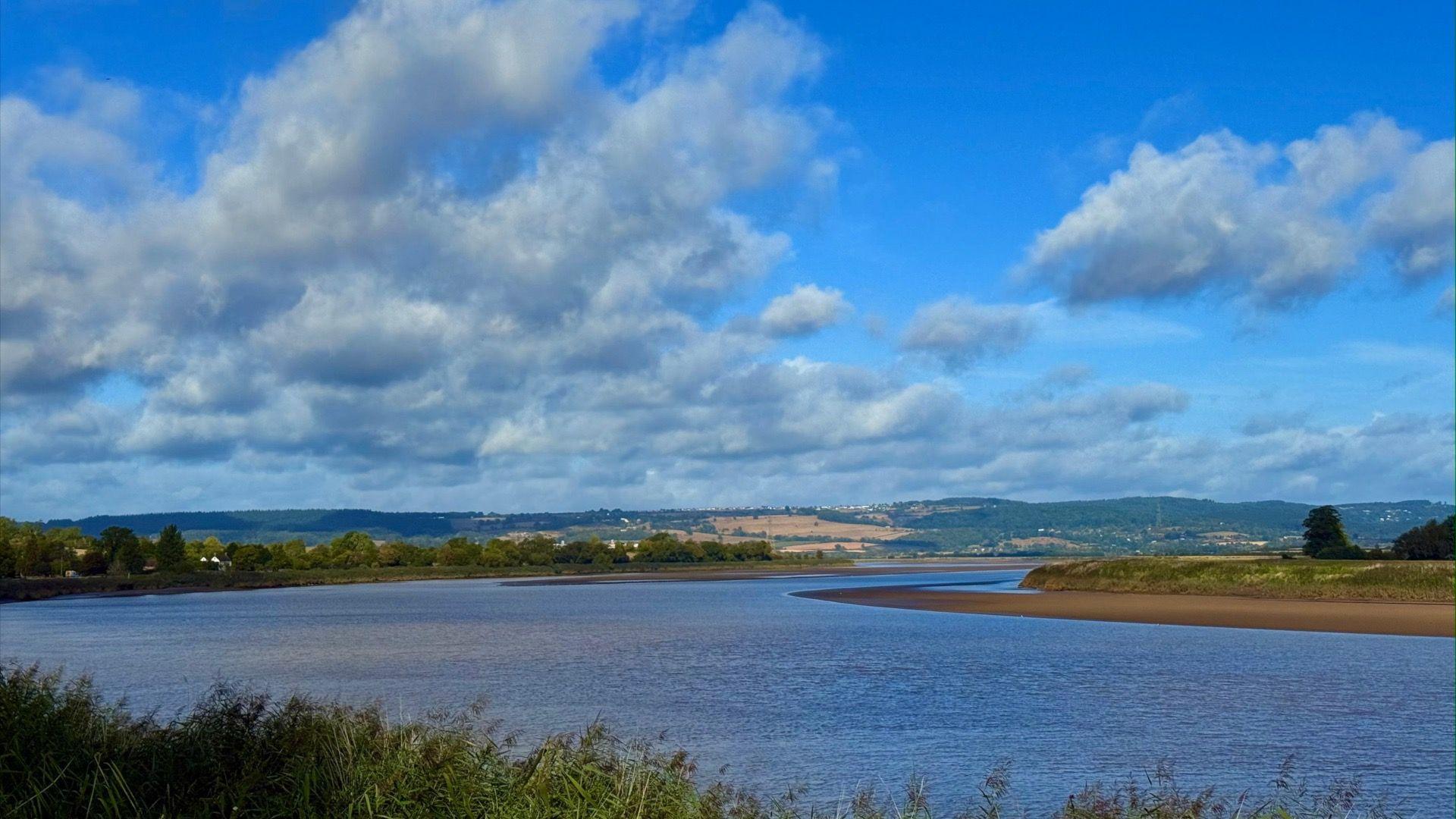 A wide river with a blue sky dotted with grey clouds above. On each bank of the river grass and trees are visible, and low hills can be seen in the distance