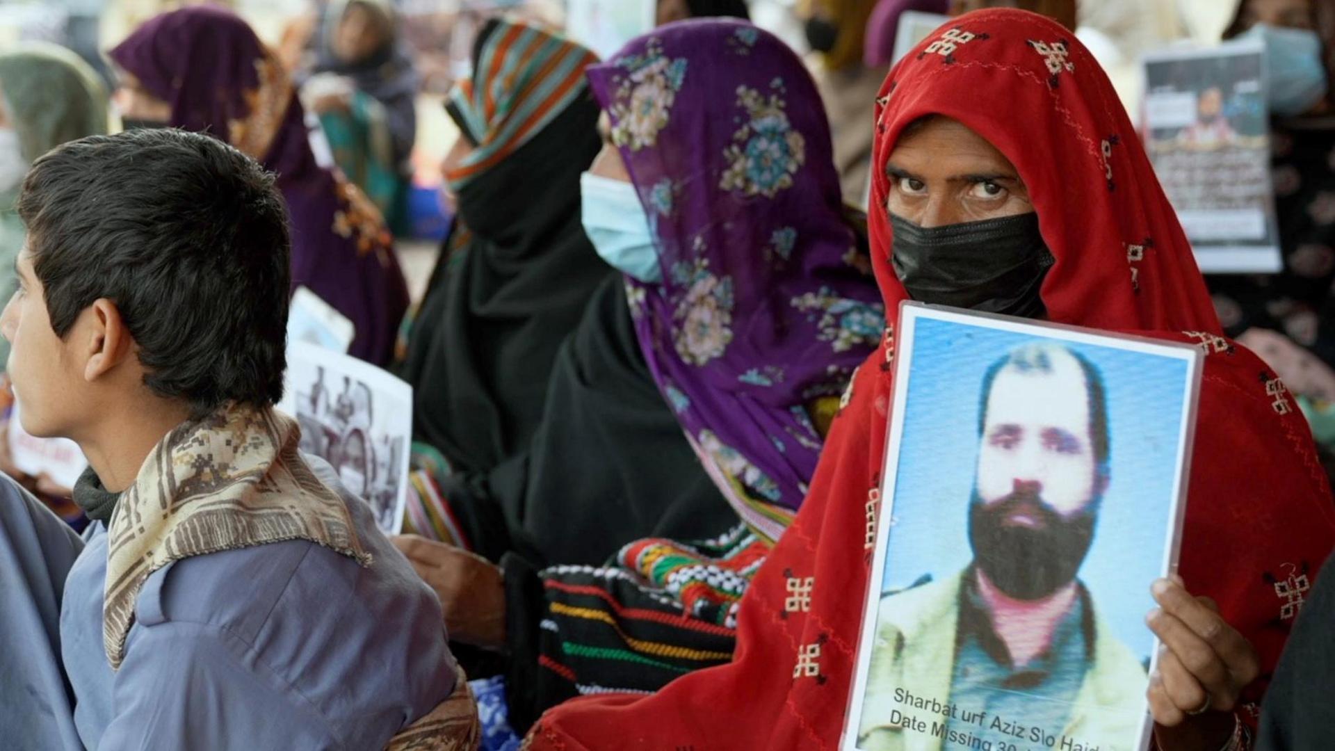 A woman with a red head covering and a mask holds up a photo of a missing relative - she is sitting in a row of women, also holding photos.  