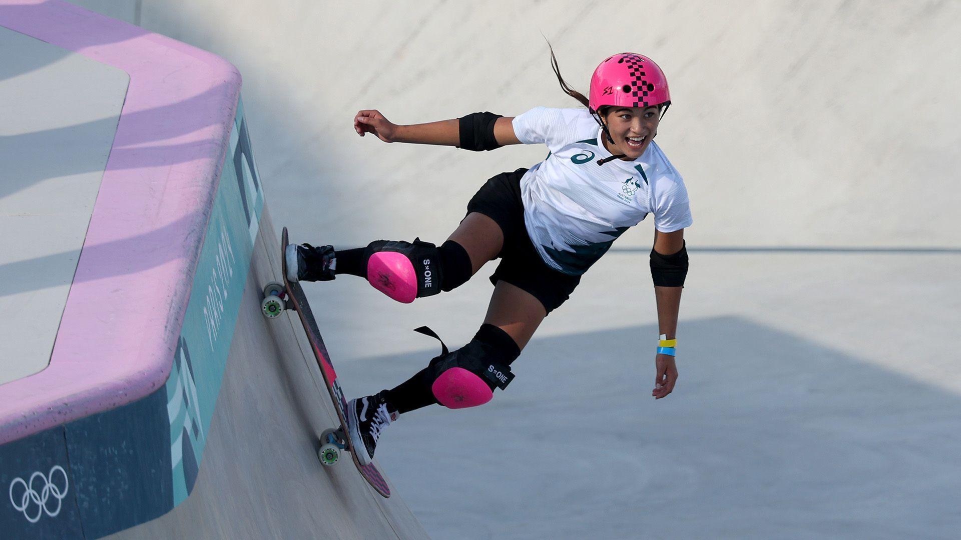 Arisa Trew rides her skateboard on a ramp. She is wearing a white top and black shorts with a bright pink helmet and kneepads. Her hair is flying behind her out of the helmet as she goes down the ramp. She is smiling and looks a mixture of shocked and happy
