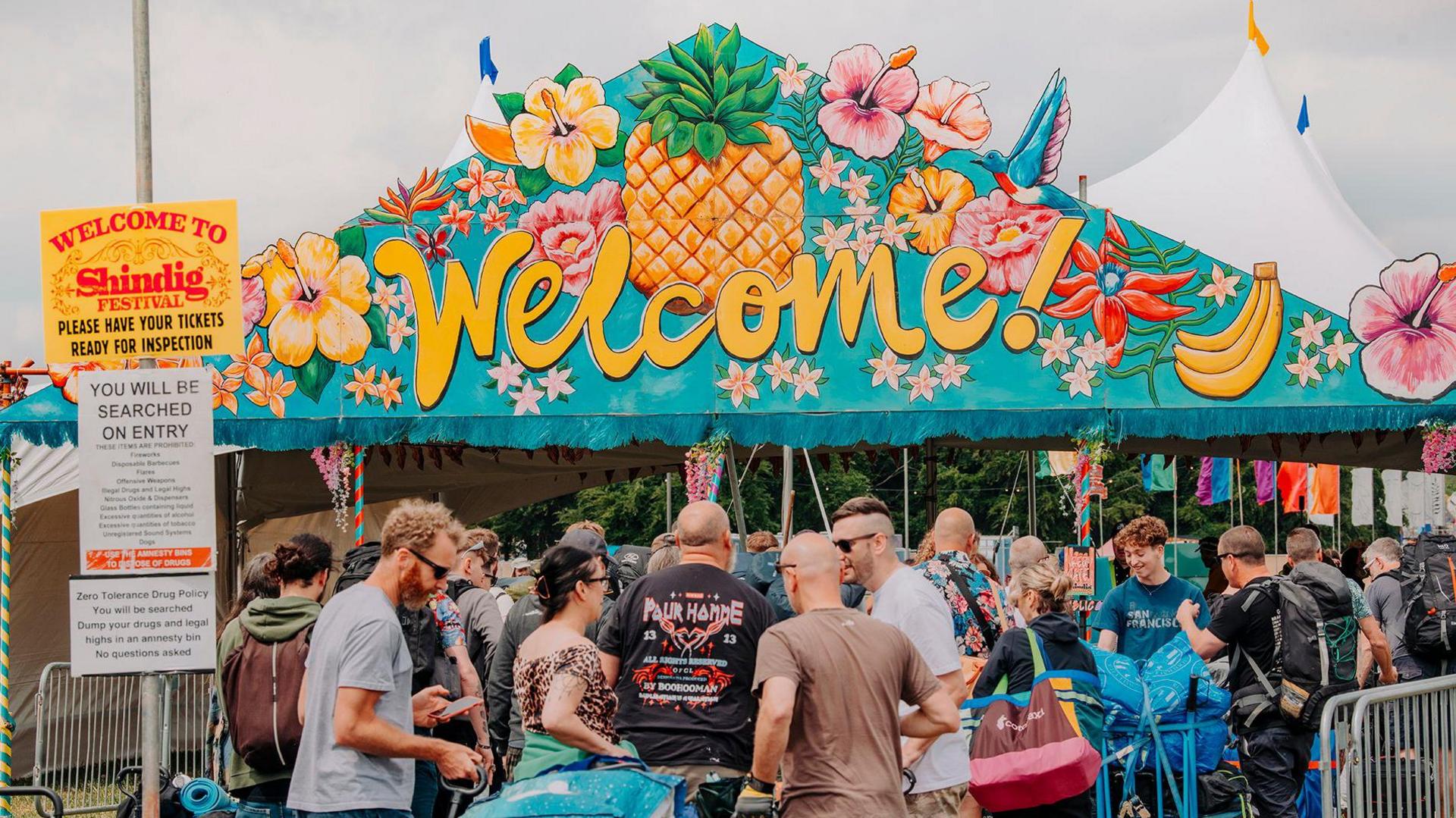 A crowd of people with camping equipment at a big welcome area, which includes a huge brightly painted sign decorated with painted fruit, birds and flowers