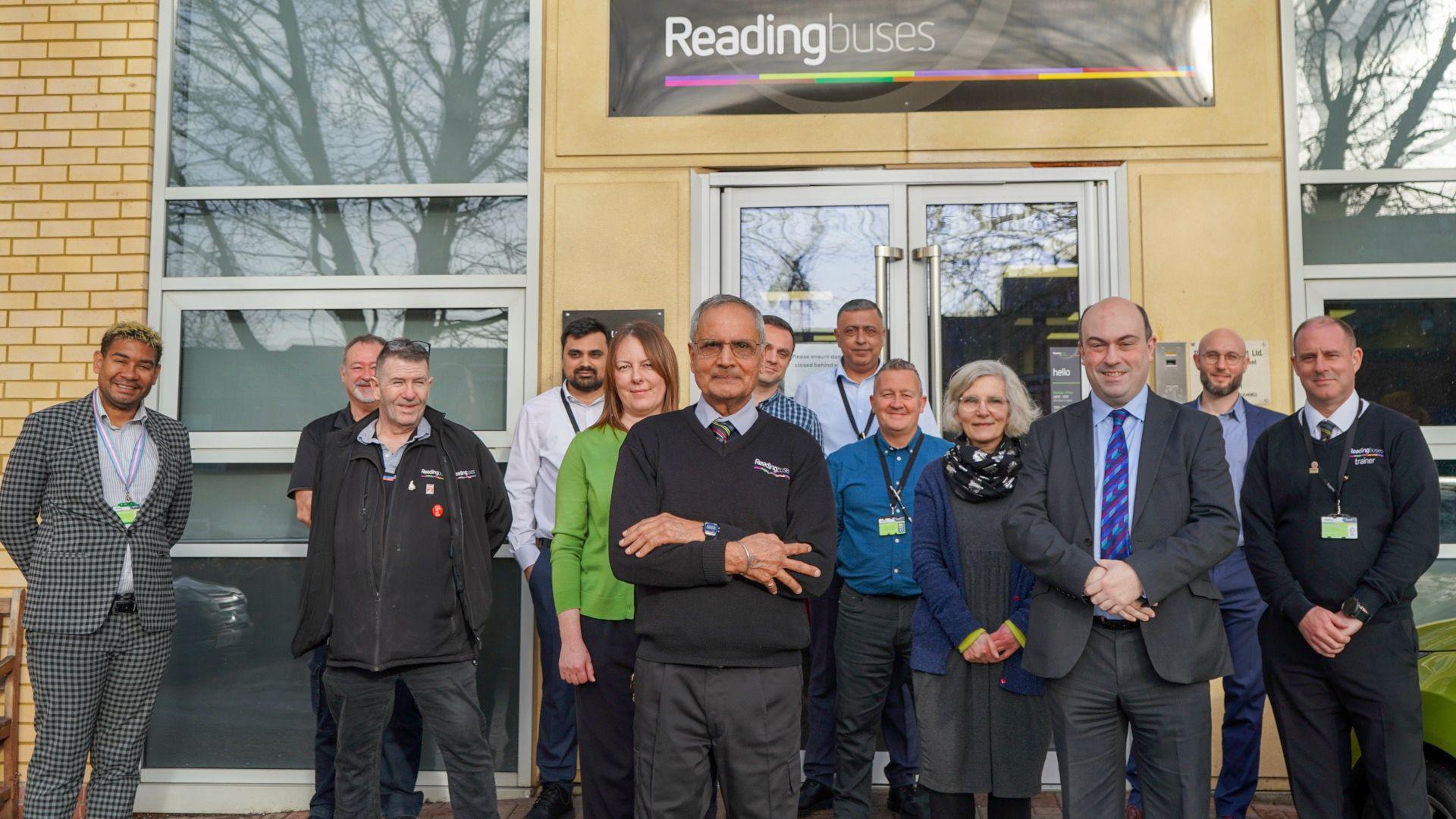 Mr Singh stands outside the Reading Buses office building surrounded by his colleagues. The group are all looking at the camera, with Mr Singh standing in the centre wearing his uniform - a blue collared shirt, tie and black jumper with grey formal trousers. There is a beige brick building behind the group with a Reading buses sign on it.