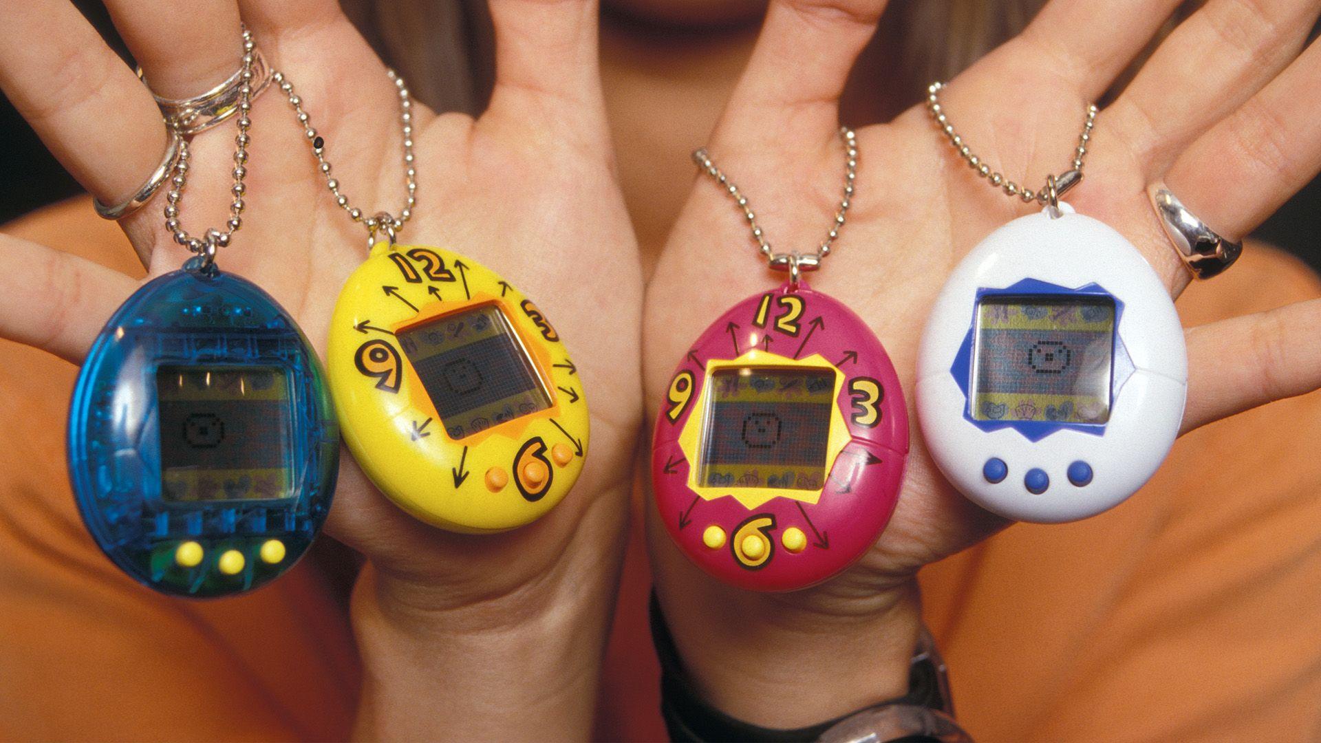 Four tamagotchi's being held by two hands. They are all different colours. From left to right they are blue, yellow, pink and white.