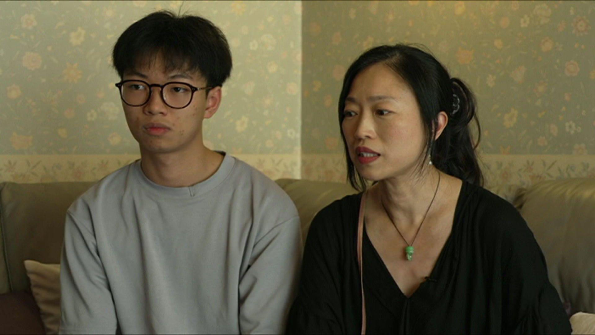 A teenage boy sits on a sofa next to his mother. He is wearing black glasses and a grey jumper. She has her hair tied back and wears a black shirt and a green pendant.