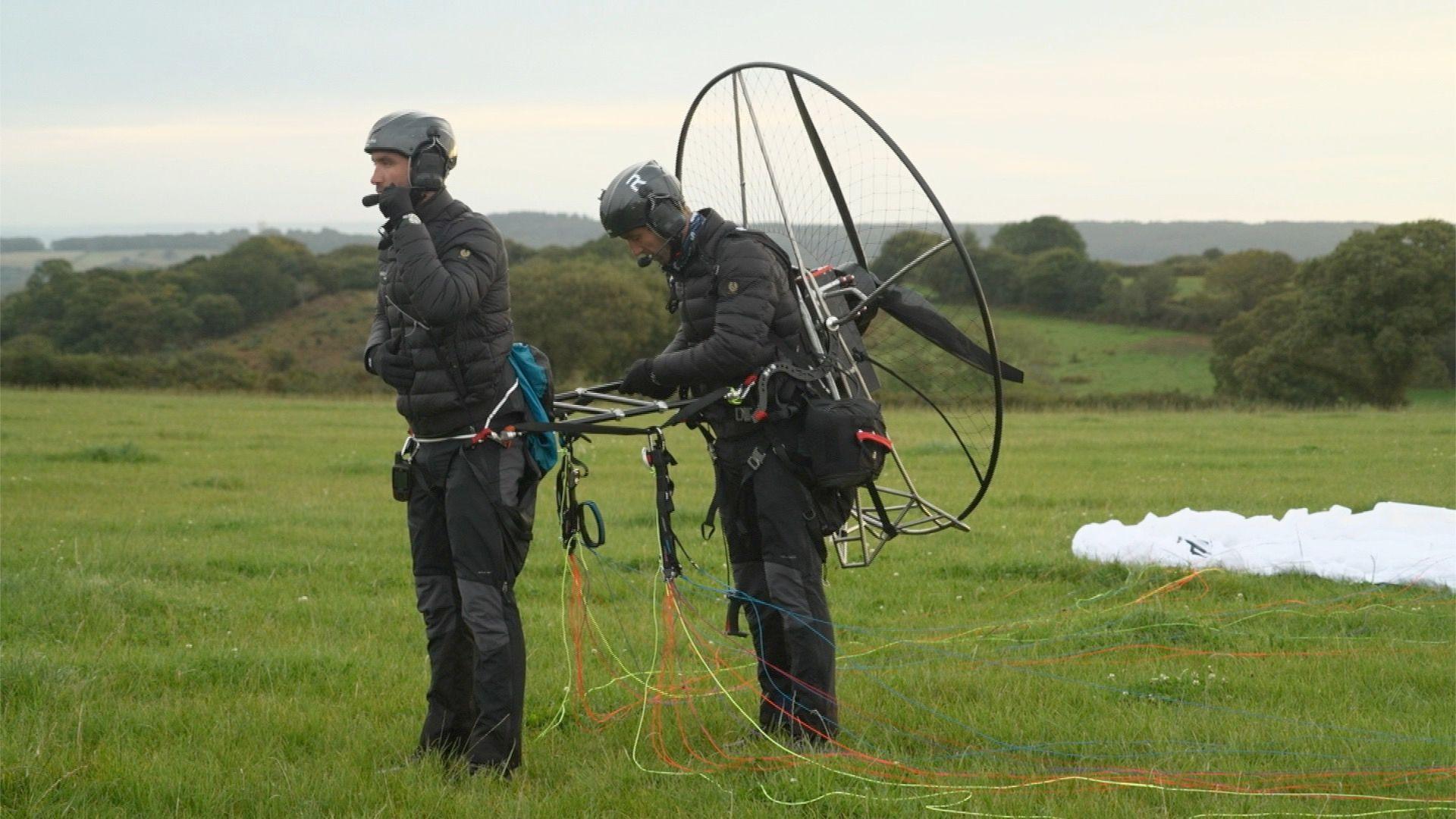 Ross and Hugo Turner preparing for a flight. They are wearing crash helmets and in black outwear, standing in a field, attached to the paramotor frame, which has a large propeller with a diameter of about 3ft (just under 1m)