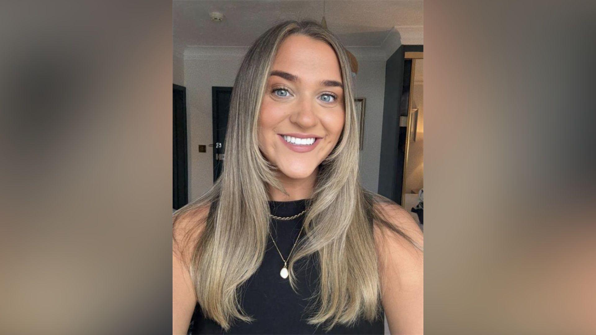 Ami Clarke in a selfie-style image. She is smiling at the camera and has long blonde hair. She is wearing a black tank top with a necklace.