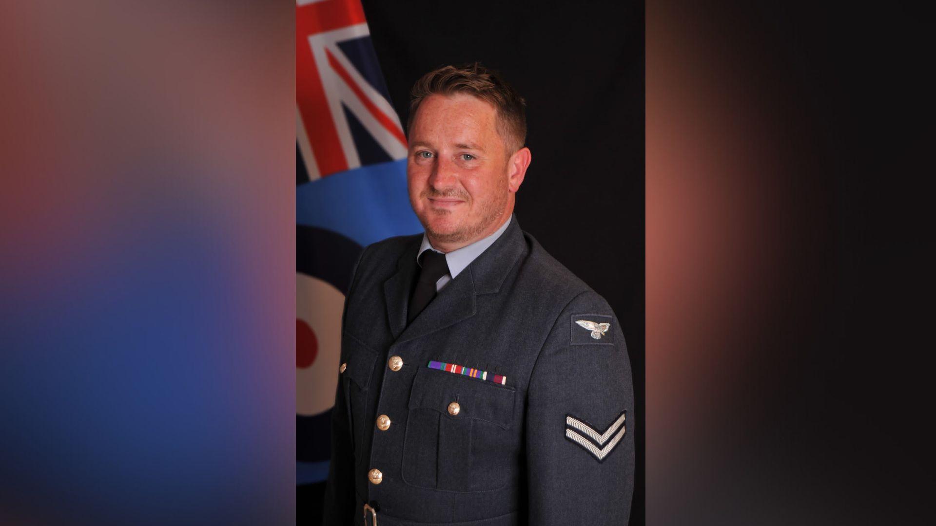 RAF veteran David Morris is dressed in a navy blue military uniform. He smiles as he looks at the camera. A Union Jack flag can be seen in the background of the photo.