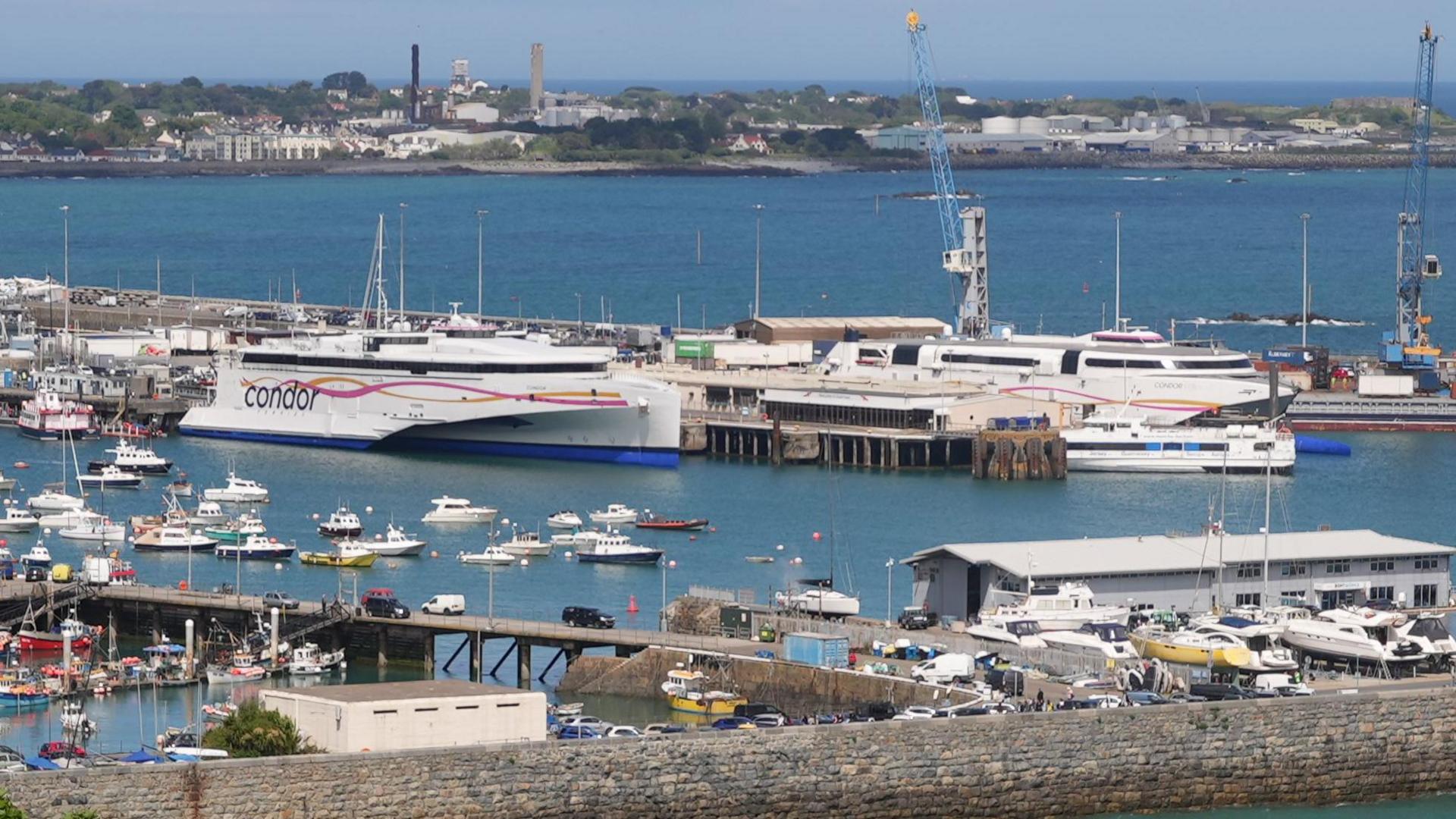 St Peter Port Harbour seen from the south with two mainly white ferries at the centre of the image. You can also see smaller vessels, harbour cranes and buildings. In the background are the two towers at the island's power station towering over buildings and trees.