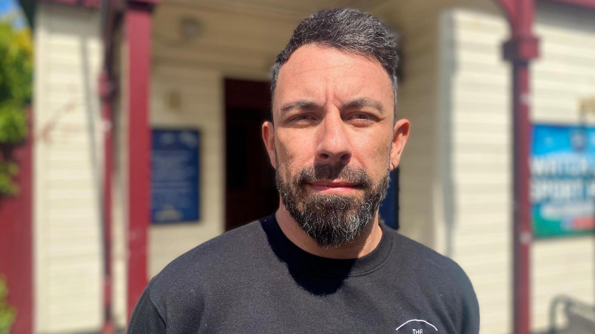 Raffael has dark hair and beard. He is standing outside his pub and is wearing a black T-shirt.