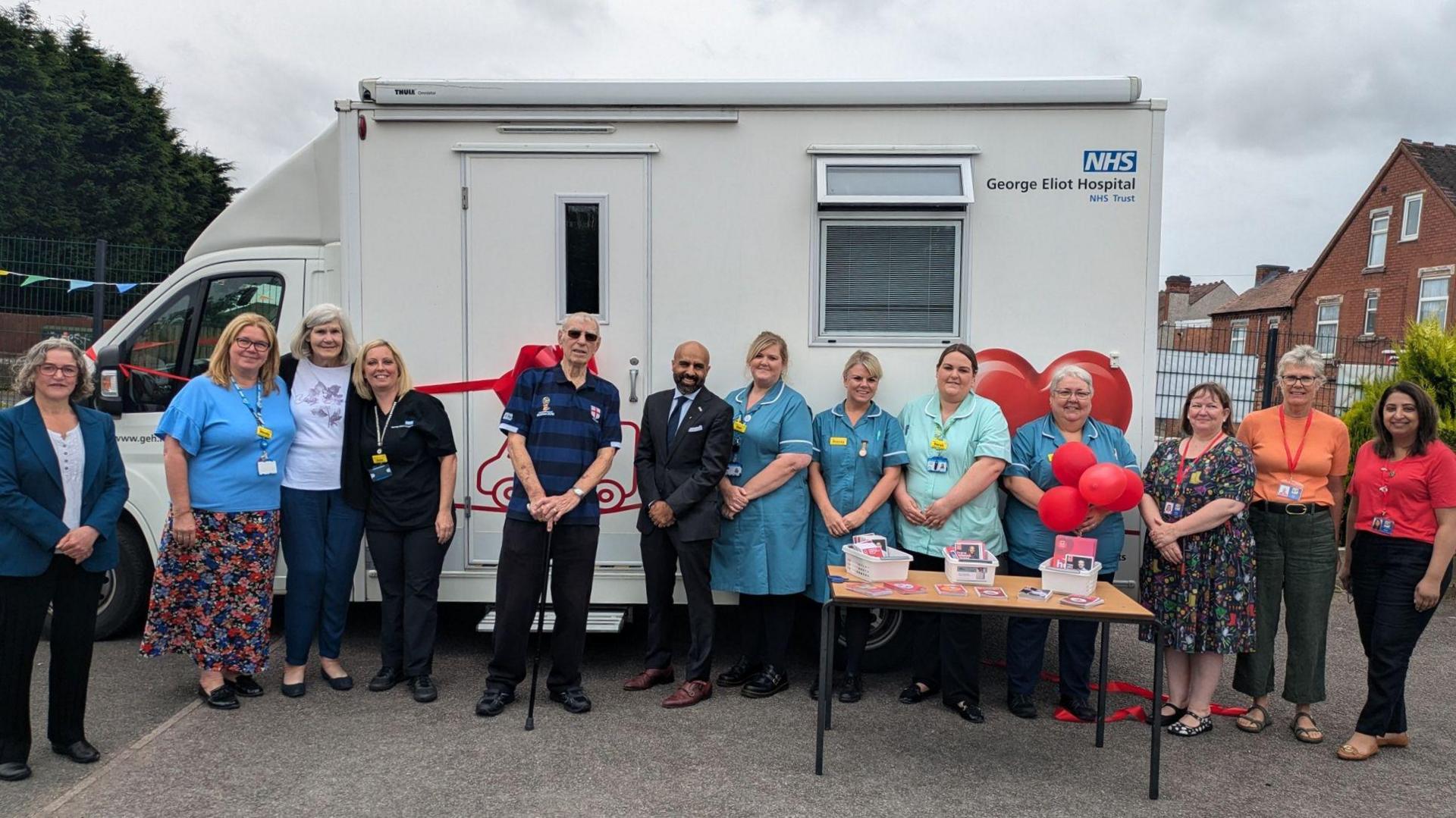 A group of people are standing in front of a white van with a red ribbon tied around it. It reads "George Eliot Hospital NHS Trust". There is a table in front of them with red balloons and leaflets.