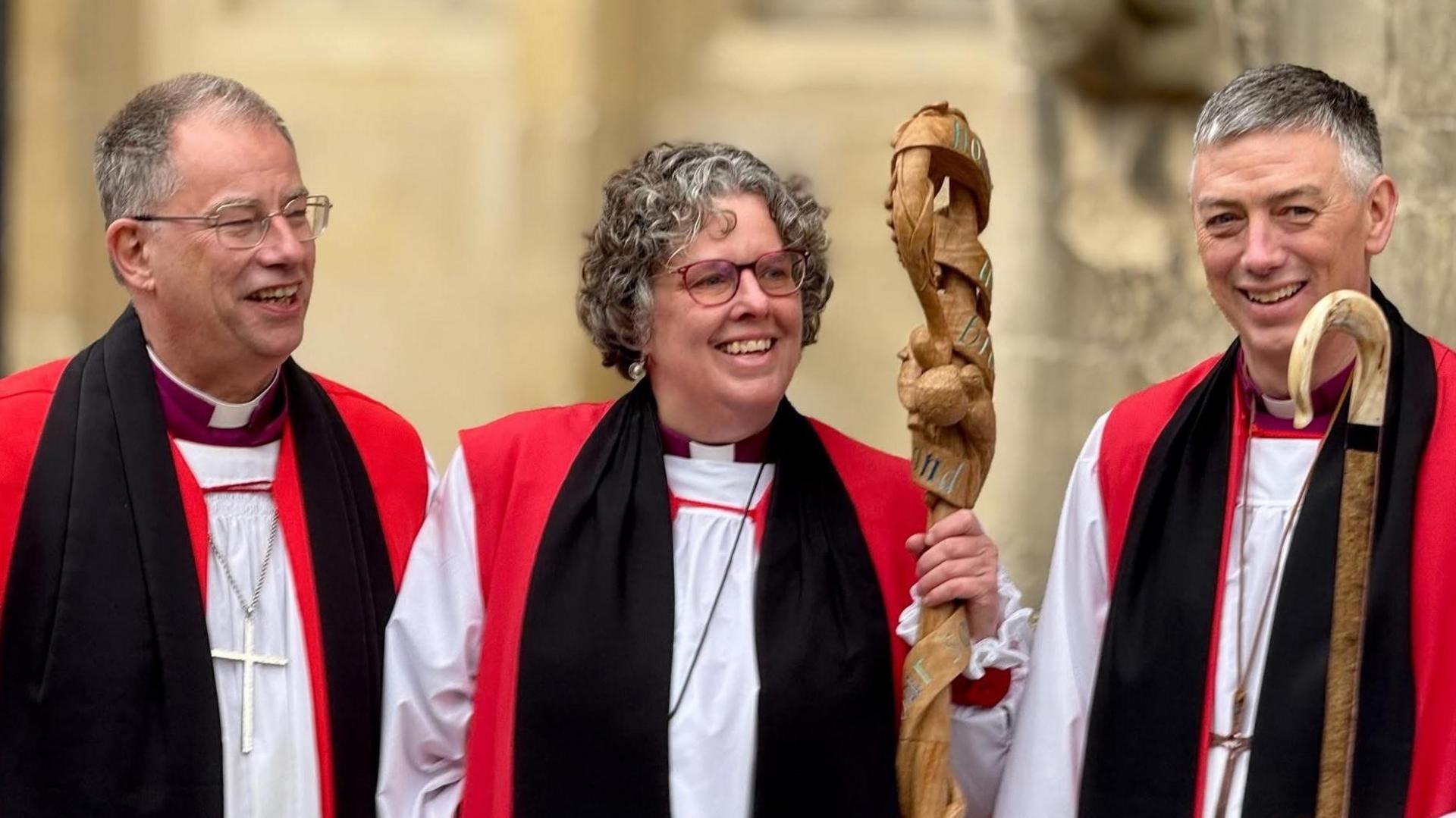 Three people are standing in the picture. The one on the right is the new Bishop of Buckingham. They are wearing a red and white robe, with a black like scarf around them. They all are wearing a cross around their necks. 