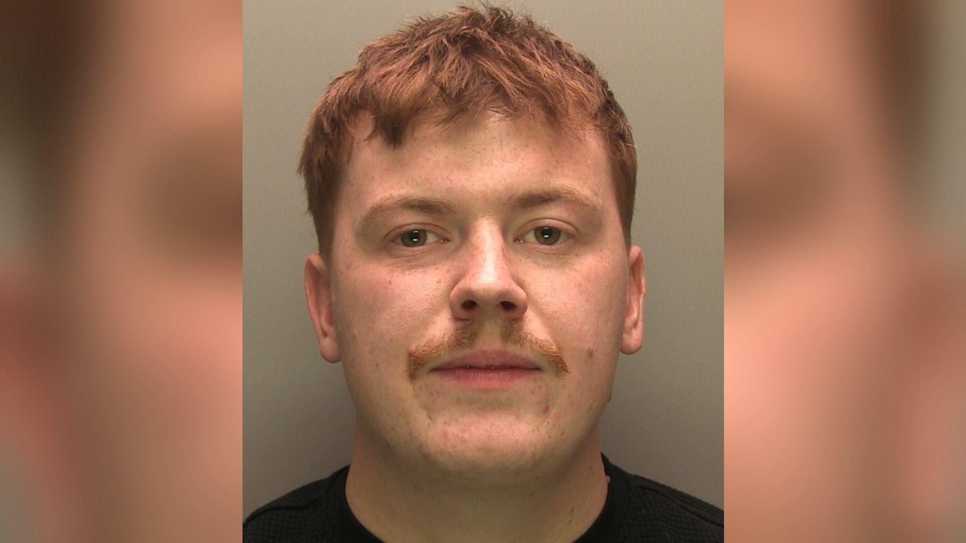 A police custody image of Brandon Barker. He has short, cropped red hair, freckled skin and a small moustache. He is staring into the camera and is wearing a black t-shirt.