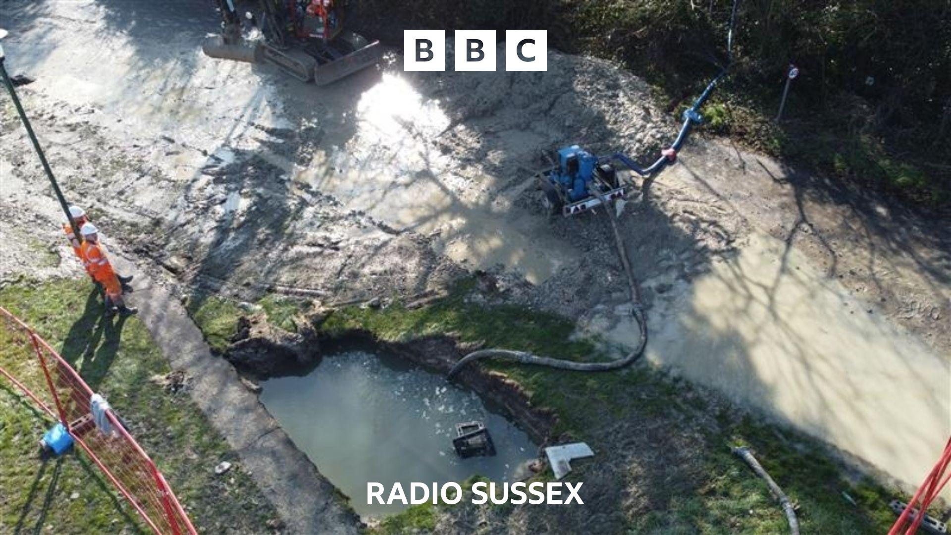 Aerial photo of the burst water main. A large hole filled with water can be seen in the bottom of the image. It is a couple of metres across. There is a pump to remove the water and a couple of workers stand in bright orange boiler suits, watching the pump's progress.