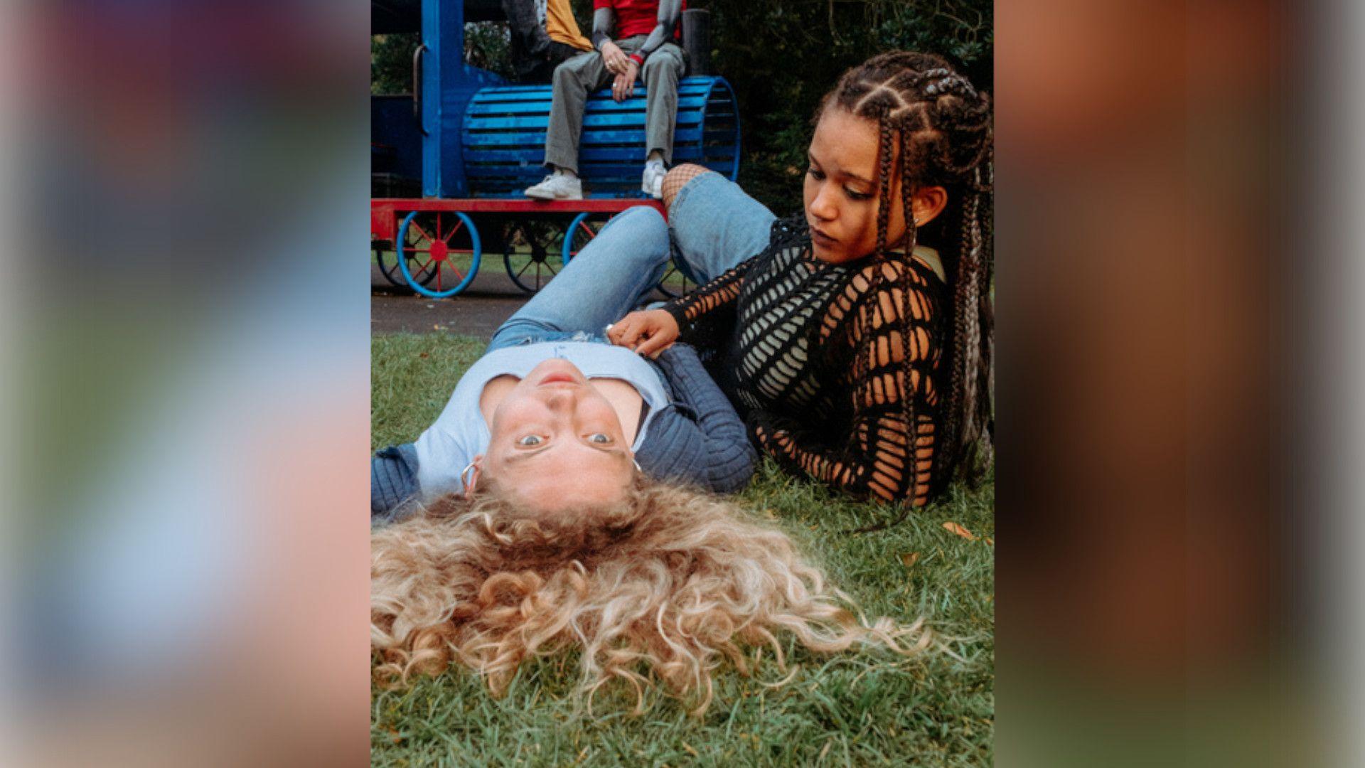 Amelia Clarkson with long curly blond hair wearing a blue top and jeans and lying on the grass of a playground. Ebony Aboagye has locs and is wearing a dark-coloured top. She is lying on the grass beside Amelia. A pair of legs in jeans sitting on a toy train is visible in the background